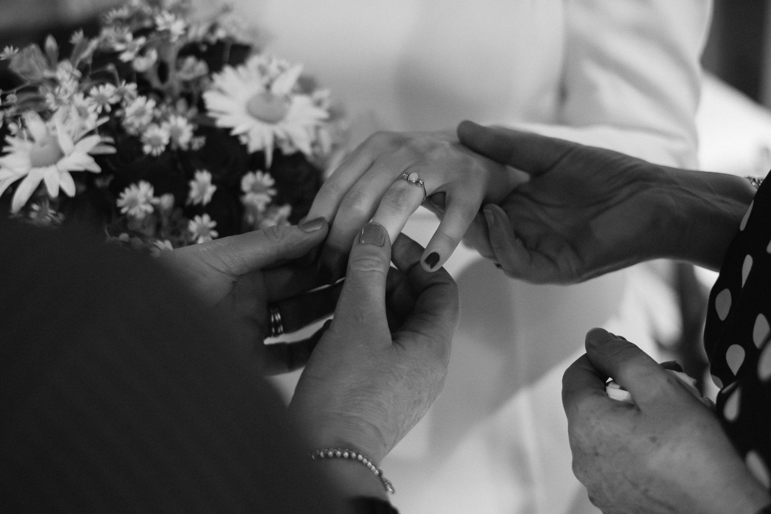 Black and white photo of hands exchanging wedding rings during a ceremony, with flowers in the background.