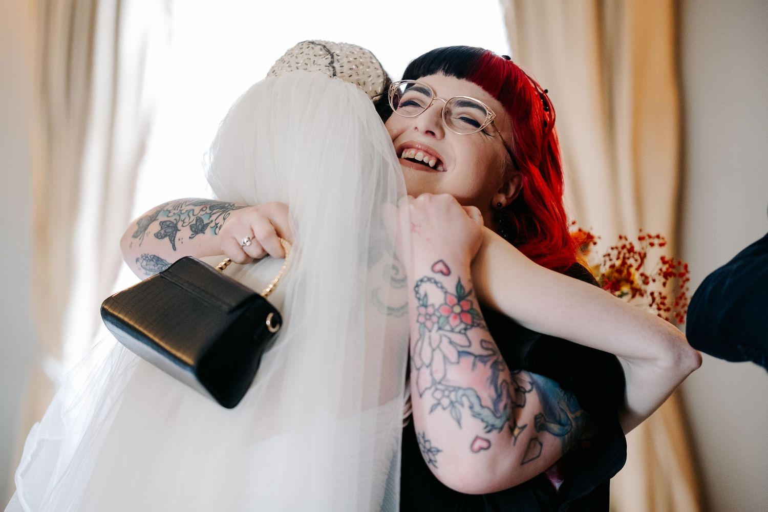 Tattooed woman with red hair hugging a bride in a white wedding dress and veil, both smiling joyfully.