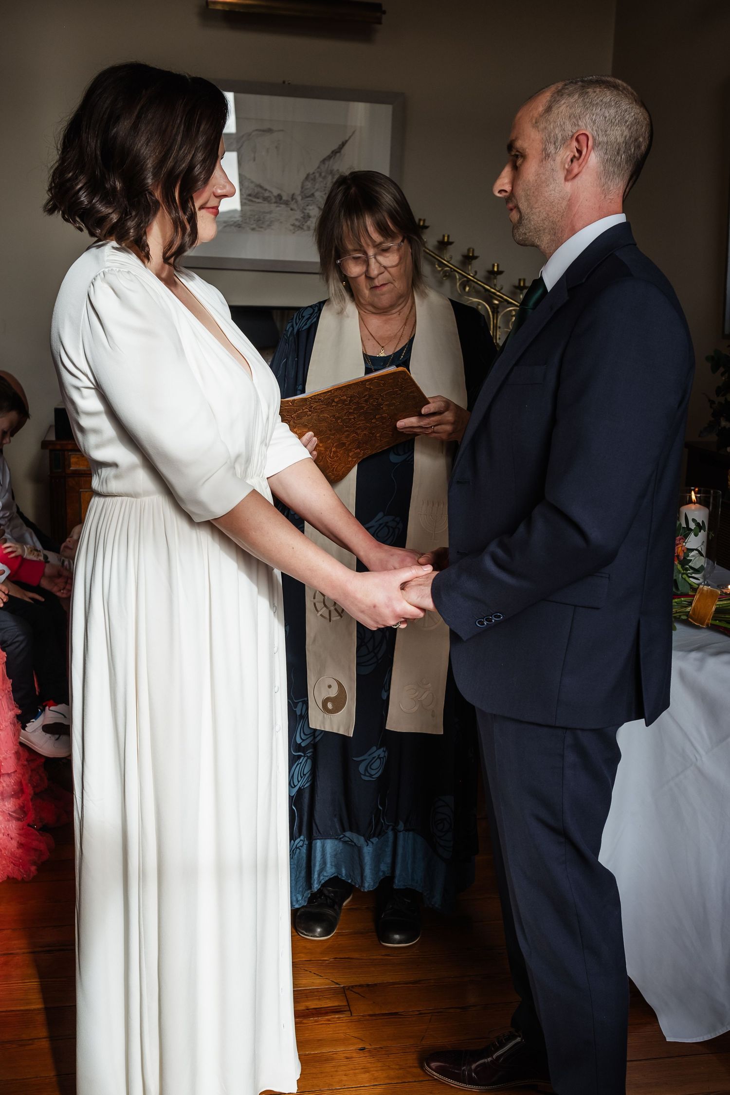 Bride and groom hold hands during intimate indoor wedding ceremony while officiant reads from book behind them.