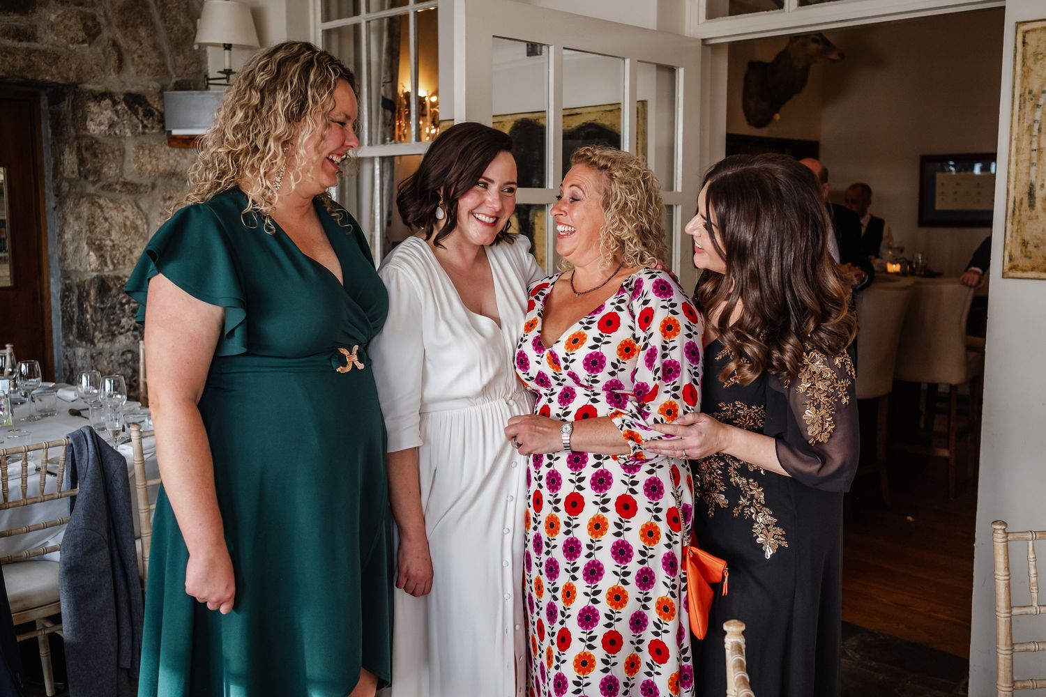 Four women laughing and socializing at a wedding reception in an elegant stone venue.