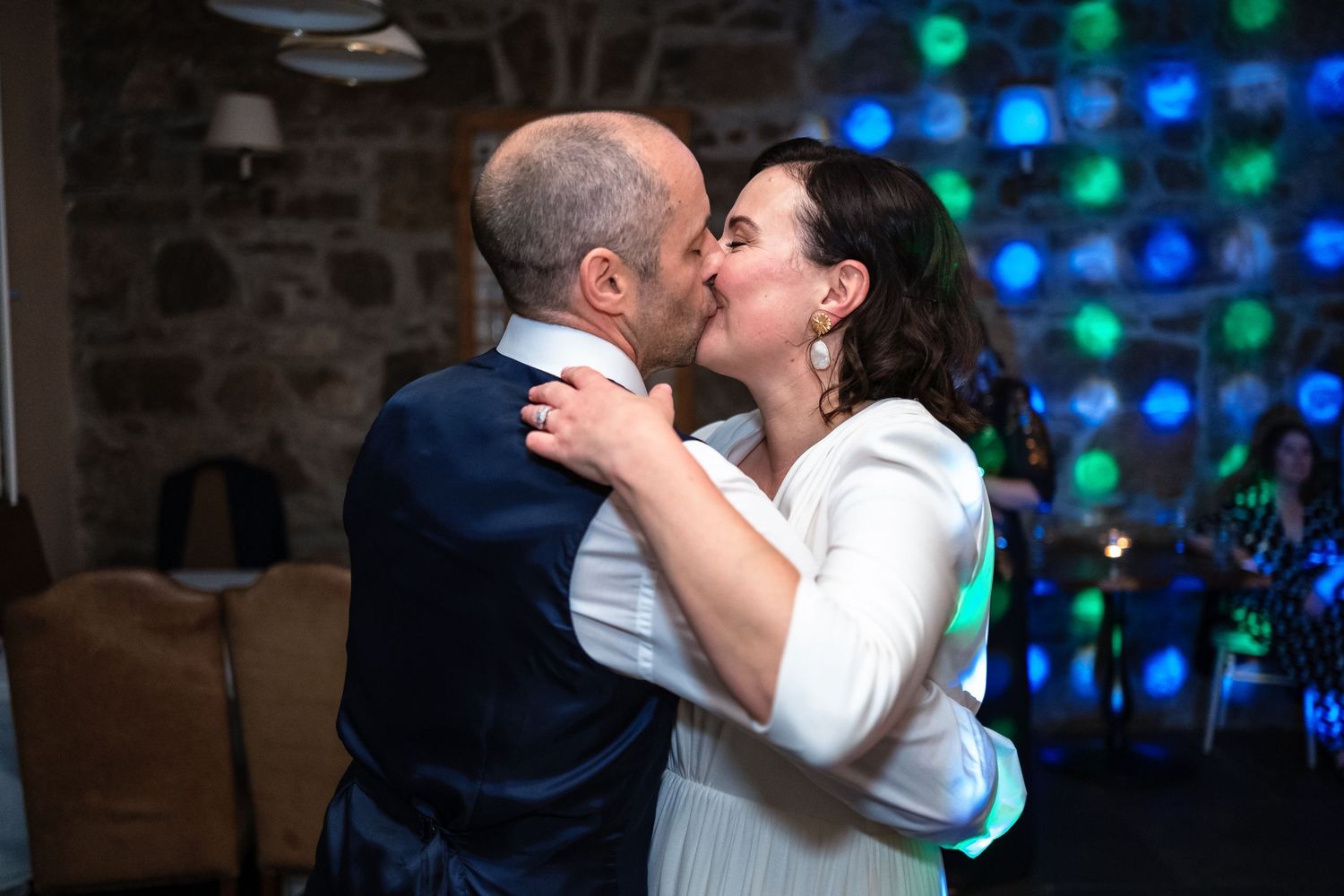 Bride and groom share a romantic first dance kiss at their wedding reception with colorful bokeh lights in background.