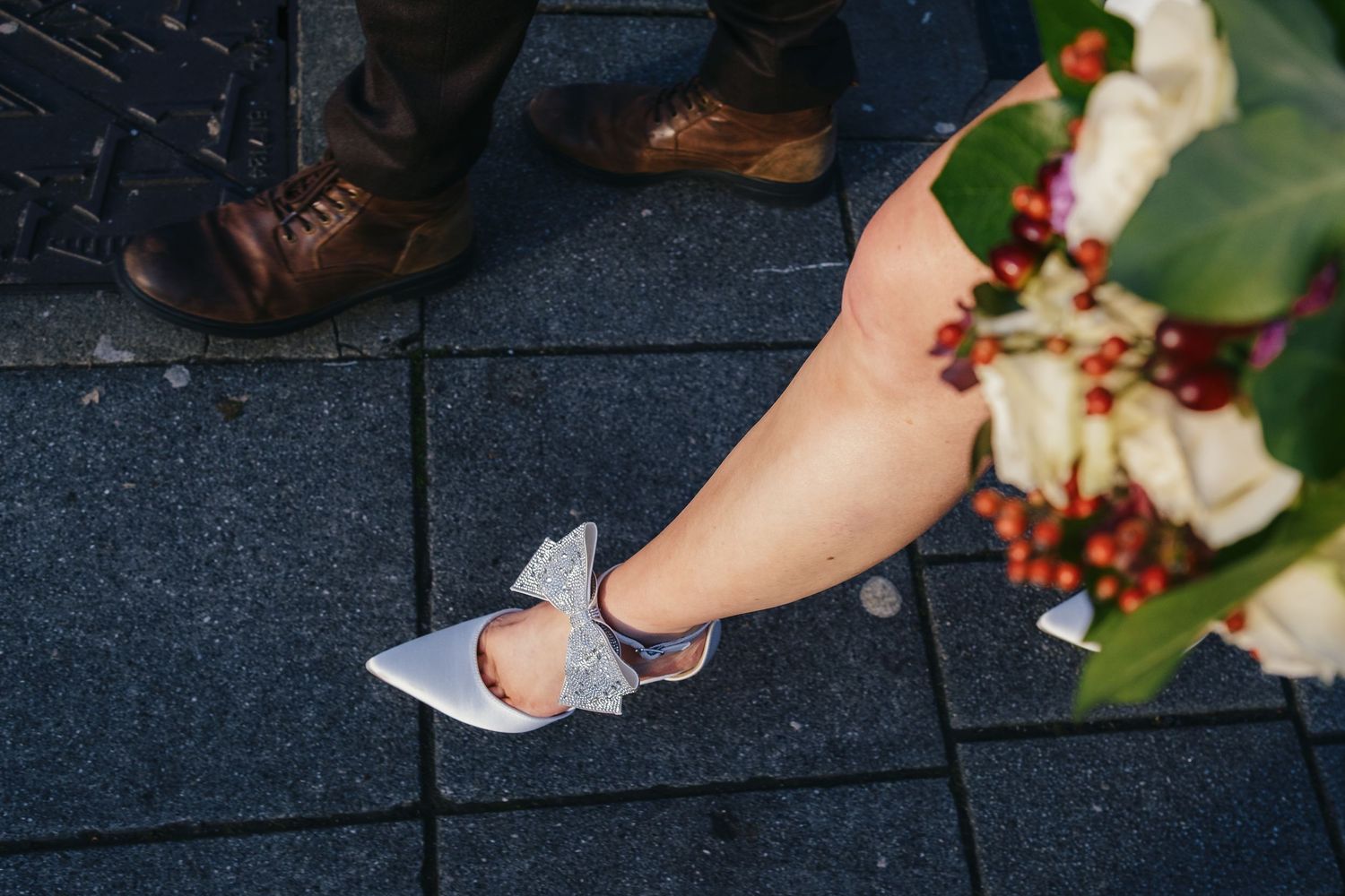 Bride wearing silver bow heels beside groom in brown dress shoes, holding a bouquet of white roses and red berries.