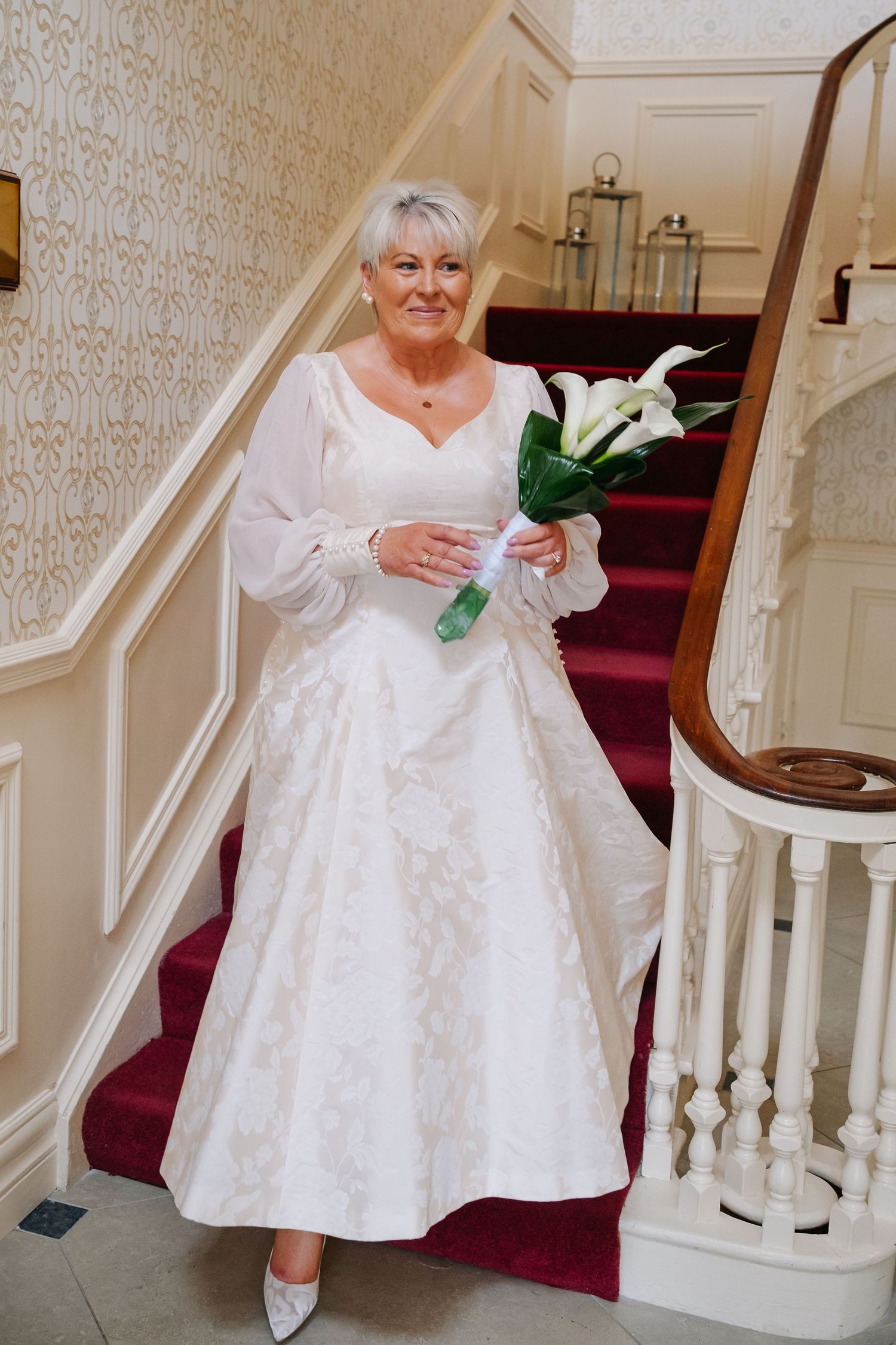 Elegant mature bride in white lace gown holding calla lily bouquet on staircase.