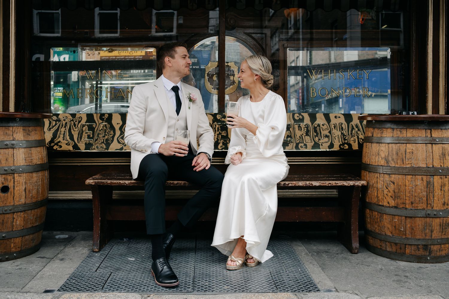 Bride and groom sitting on a bench outside a pub, sharing drinks and smiling at each other on their wedding day.
