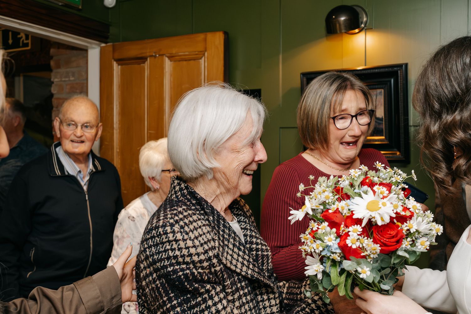 Elderly woman smiling joyfully at a celebration, receiving a bouquet of red and white flowers indoors.