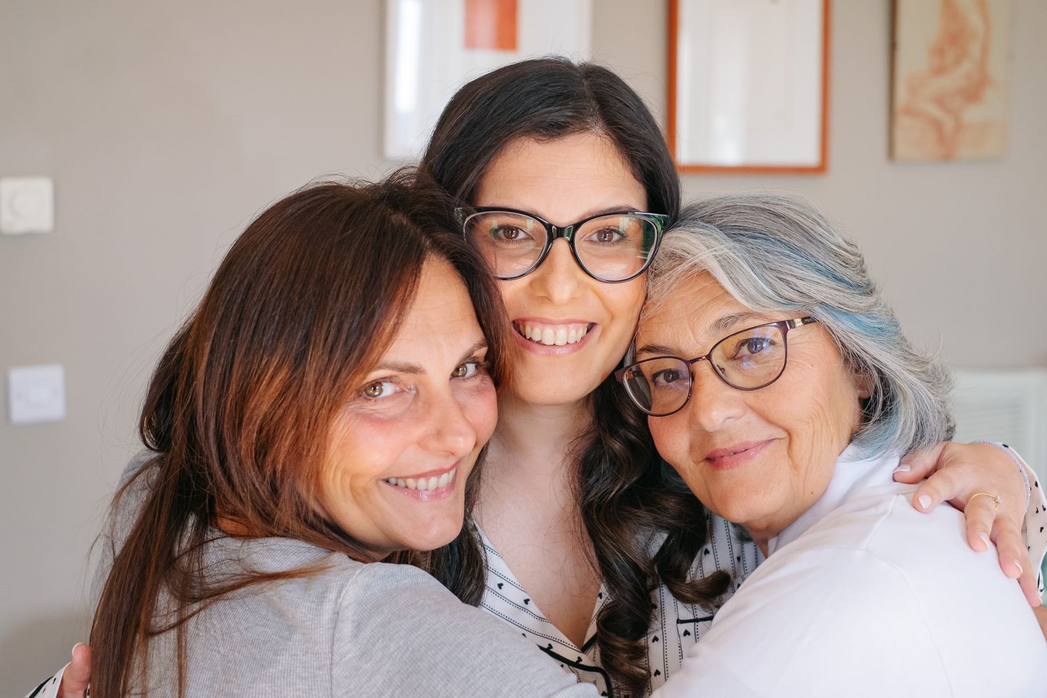 Three women of different ages smiling and hugging together in a warm, joyful family portrait indoors.