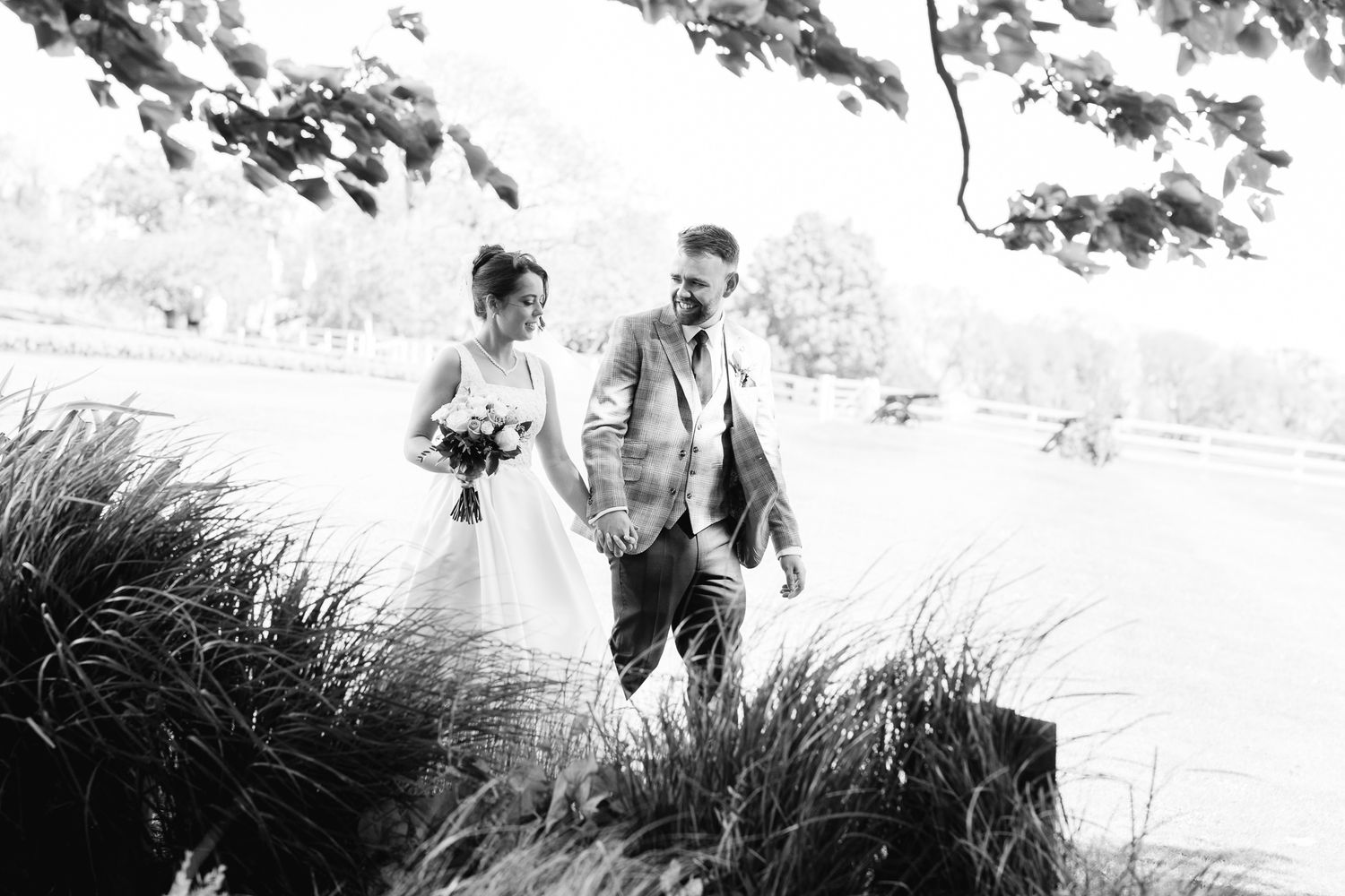 Black and white photo of a bride and groom holding hands walking through tall grass at an outdoor wedding venue.