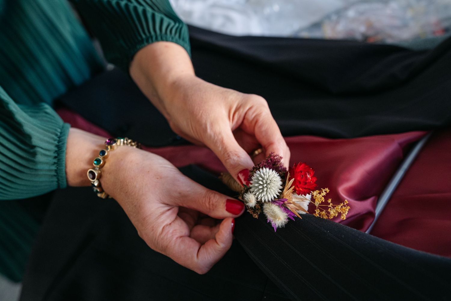 Close-up of hands with red nails and gold bracelet pinning a floral boutonniere onto dark fabric.