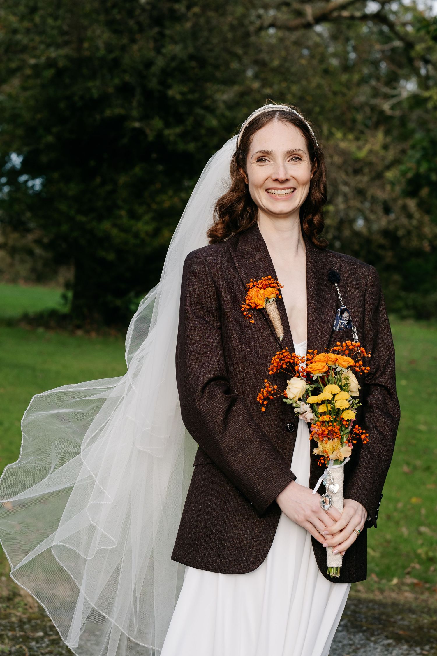 Bride wearing white dress and brown jacket holding orange and yellow autumn bouquet.