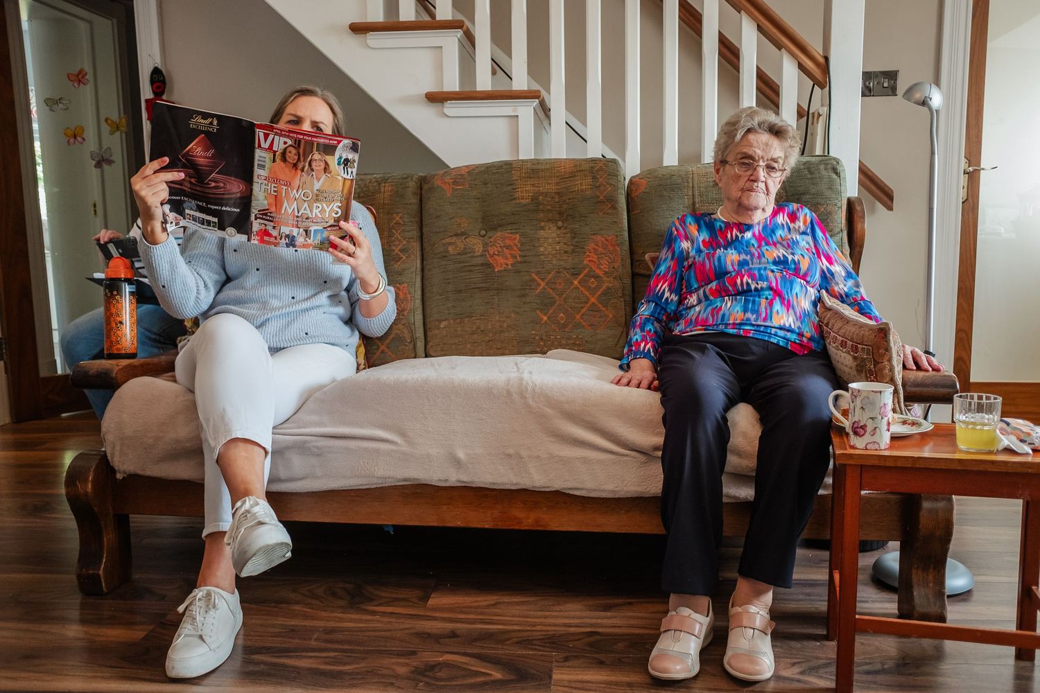 Two people sitting on brown couch beneath staircase in home interior.