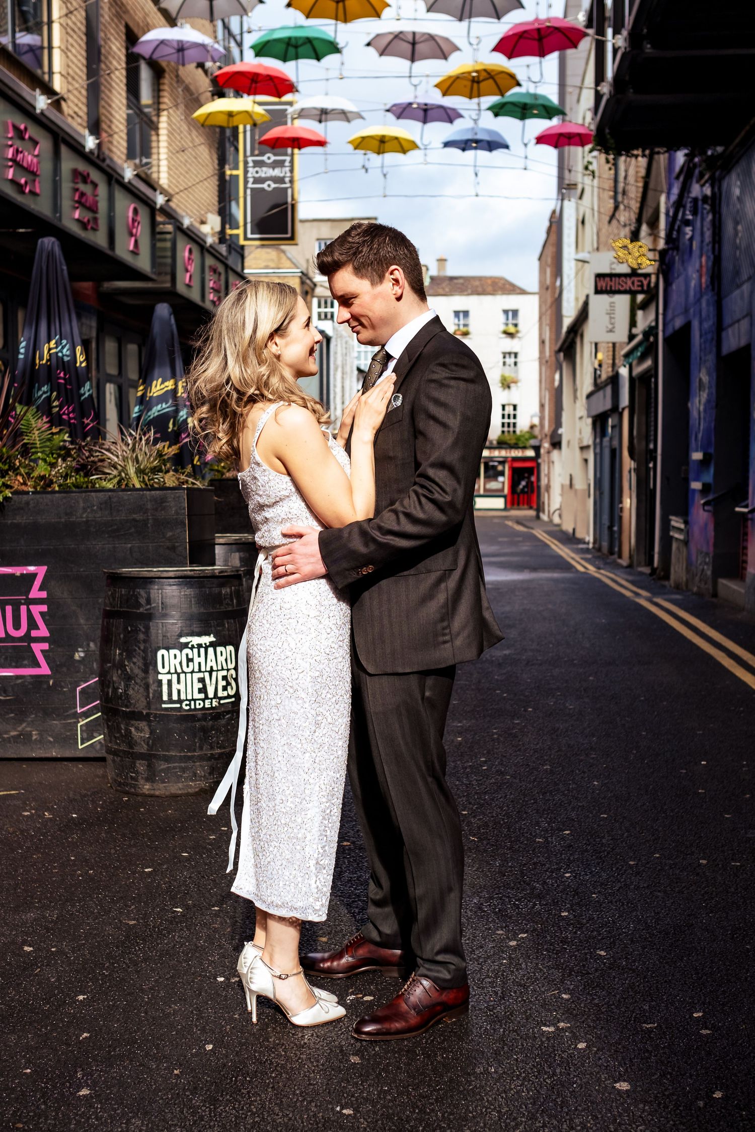 Couple in formal wear embraces under hanging umbrellas in urban alley.
