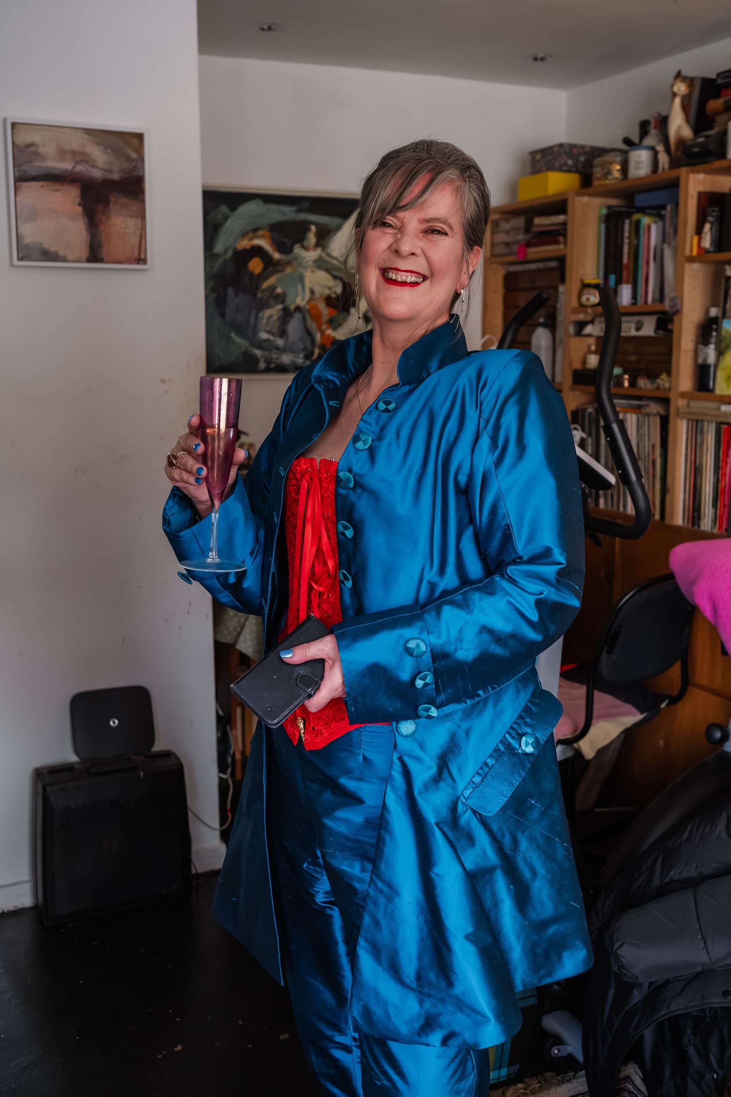 Person in bright blue silk jacket and red corset standing in front of bookshelves.