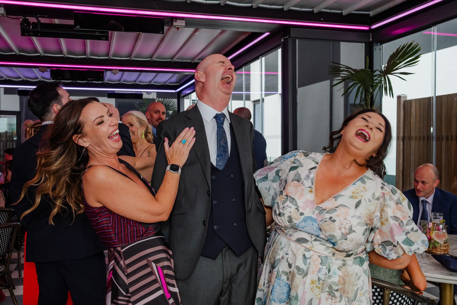 Wedding reception celebration with guests laughing and dancing under purple lighting.