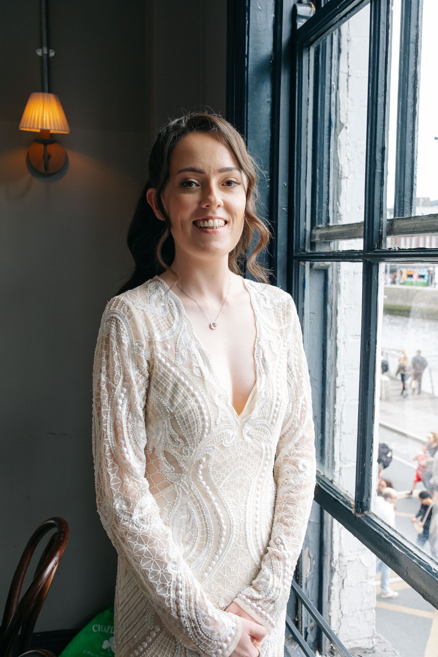 Bride in an elegant white lace wedding dress poses near windows in an indoor setting.
