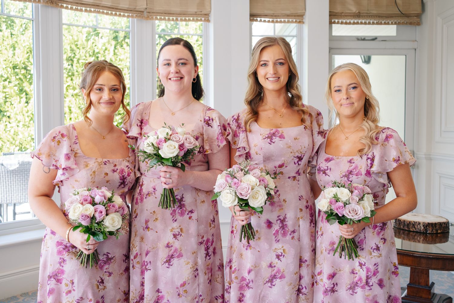 Four bridesmaids in matching pink floral dresses holding white and pink bouquets, posing together near a bright window.