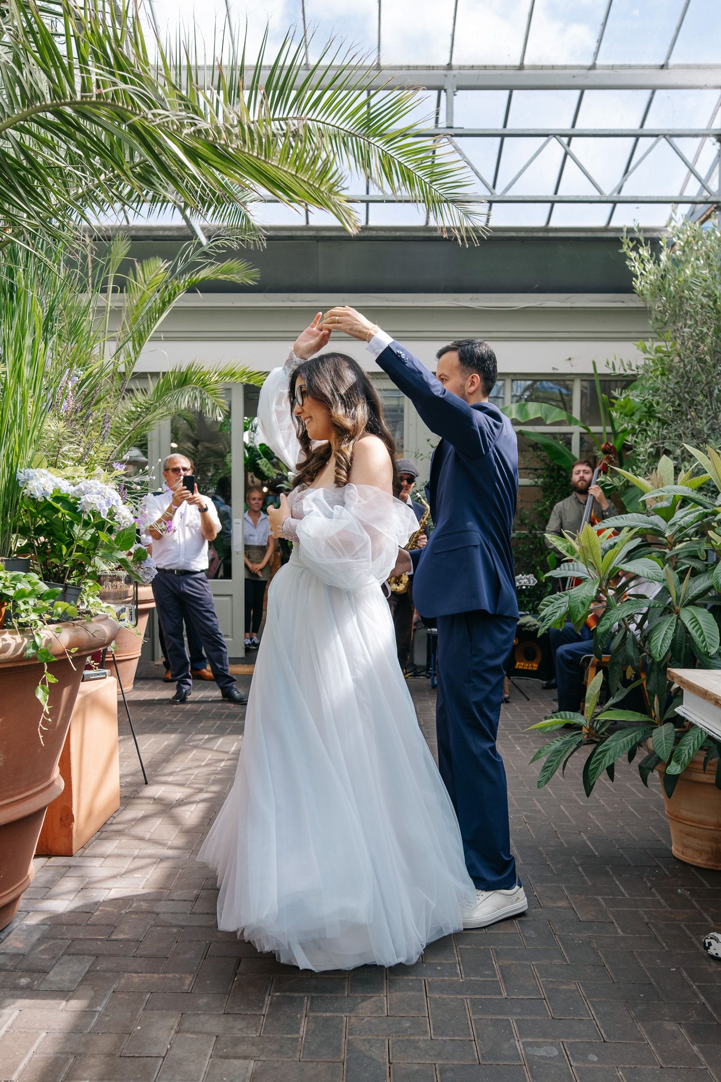 Bride and groom share first dance in a greenhouse venue, surrounded by lush tropical plants and wedding guests.