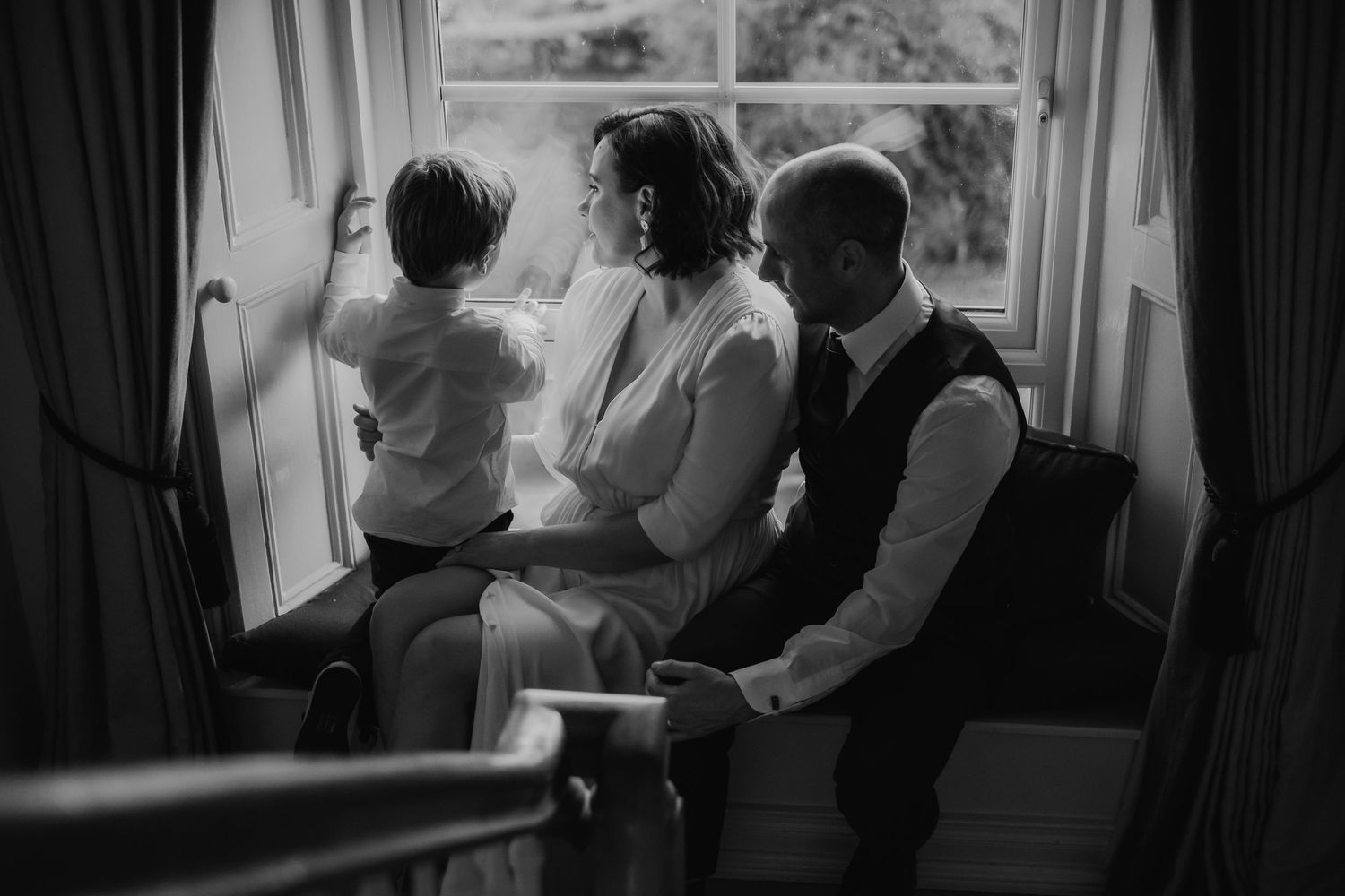Black and white photo of a couple and child sitting by a bright window, capturing an intimate family moment.
