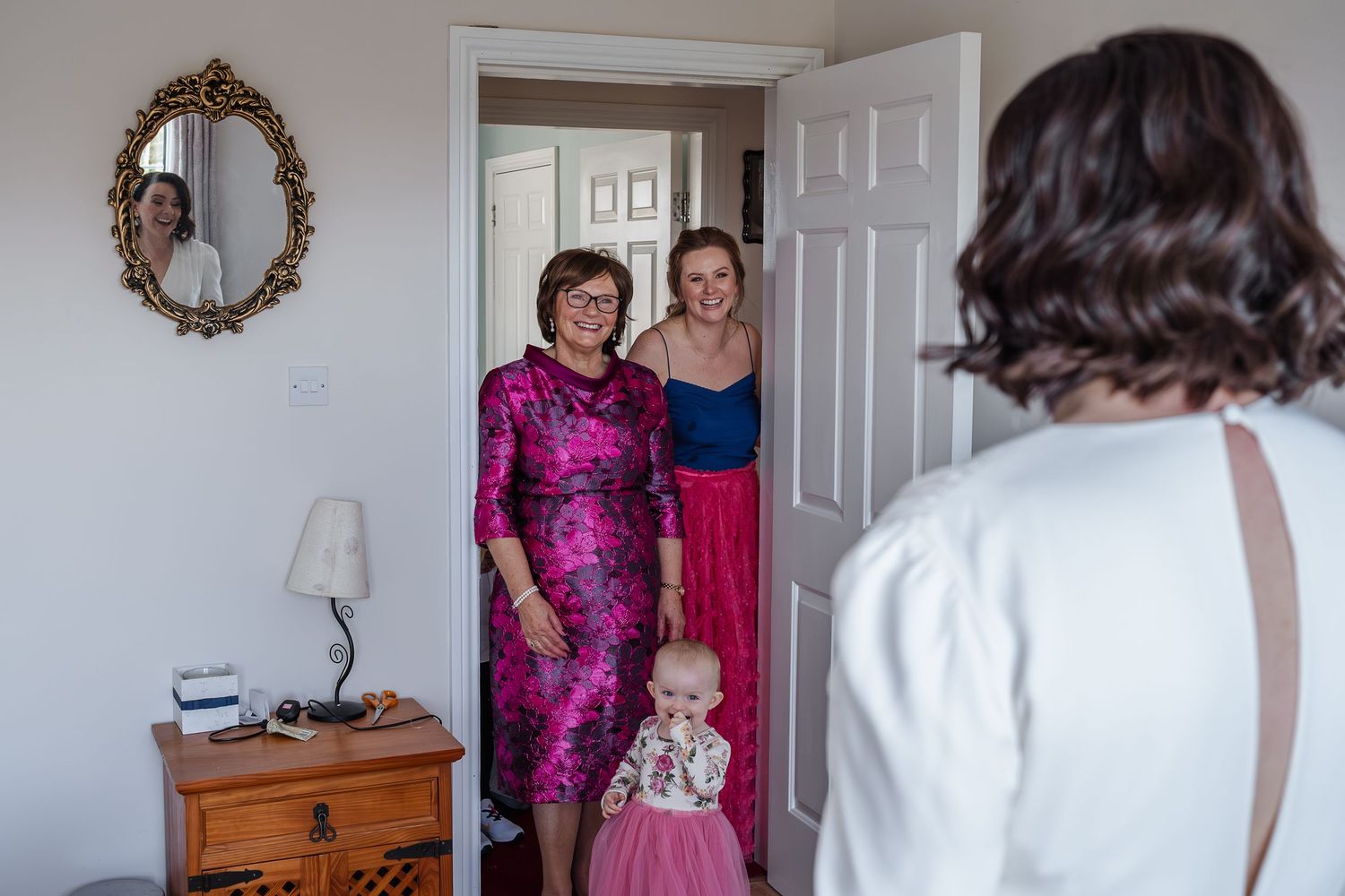 Woman in pink dress and daughter in blue gown posing in doorway with baby at wedding.