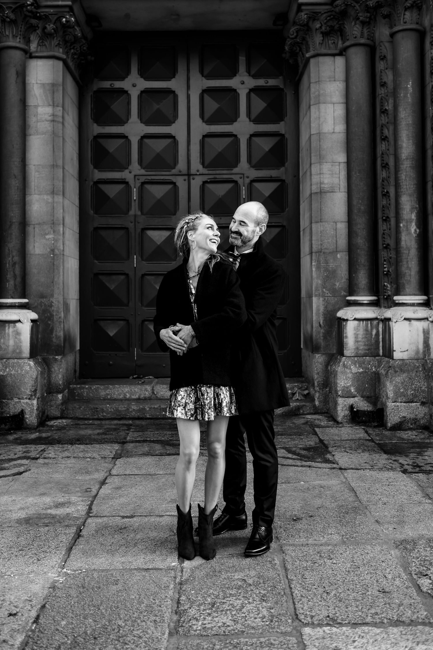 Black and white photo of a smiling couple embracing before large ornate doors.