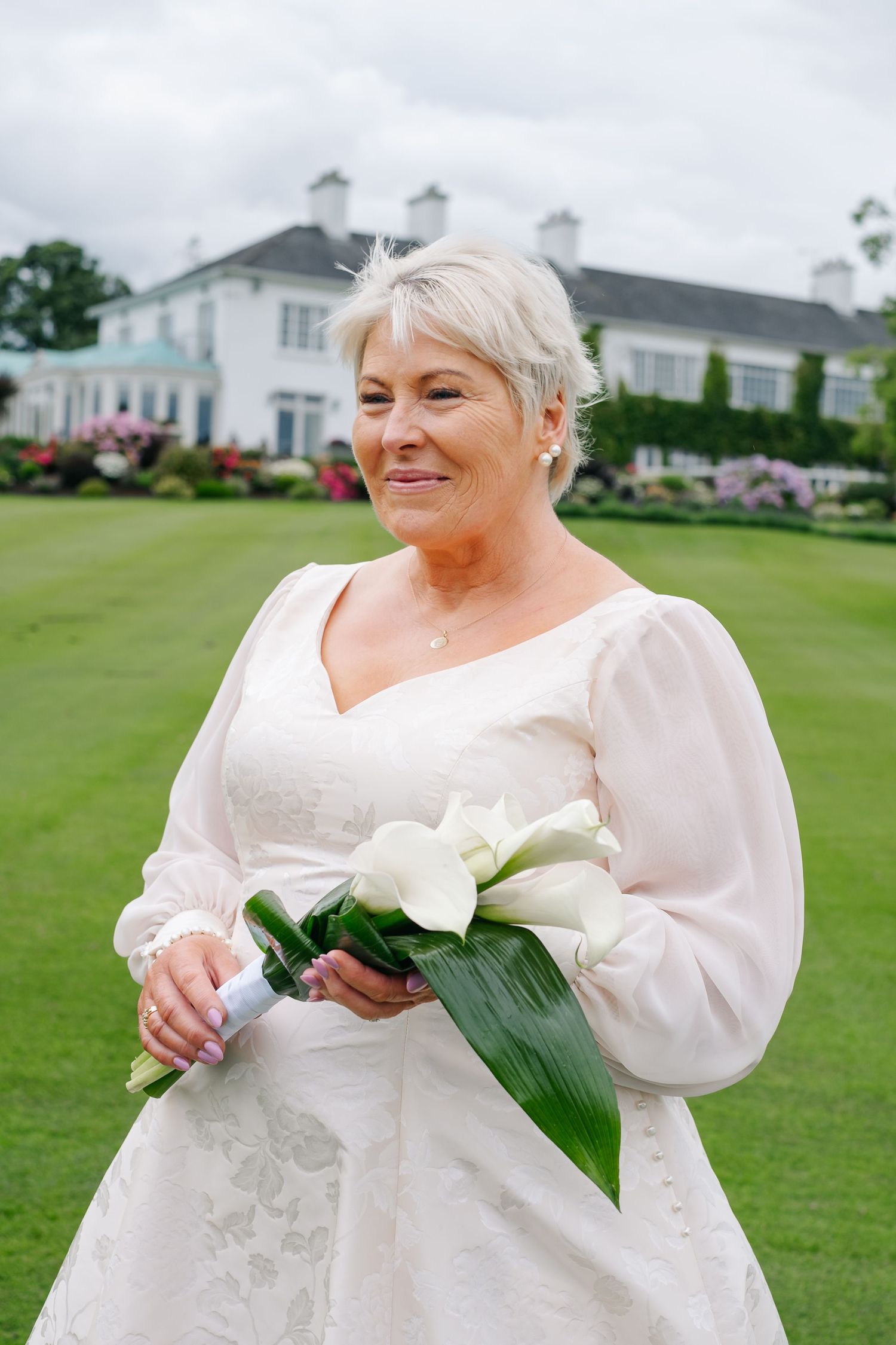 An elegant older bride in a white gown holds white calla lily bouquet outdoors at a manor estate.