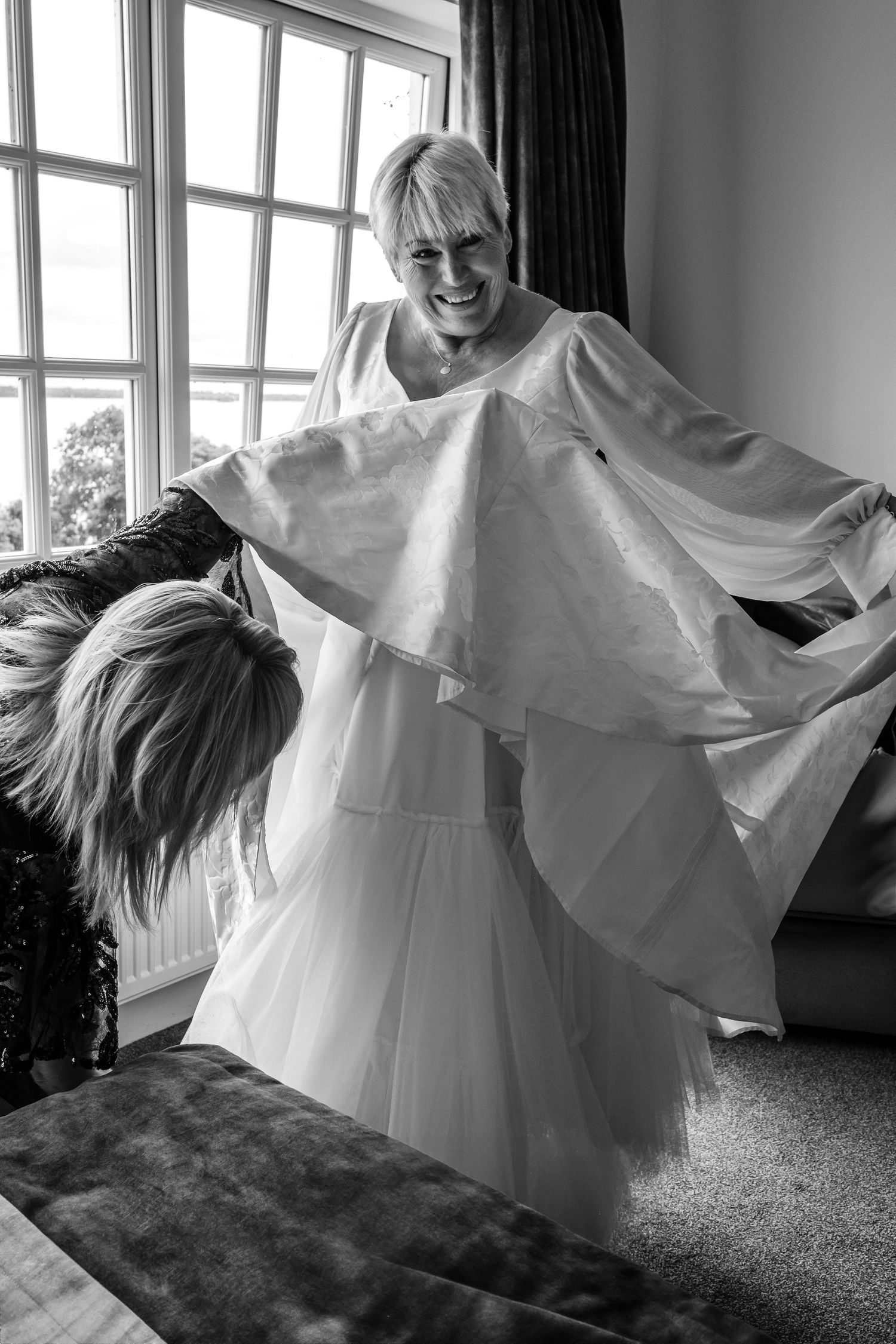 Bride smiling joyfully as assistant helps her into flowing white wedding gown.
