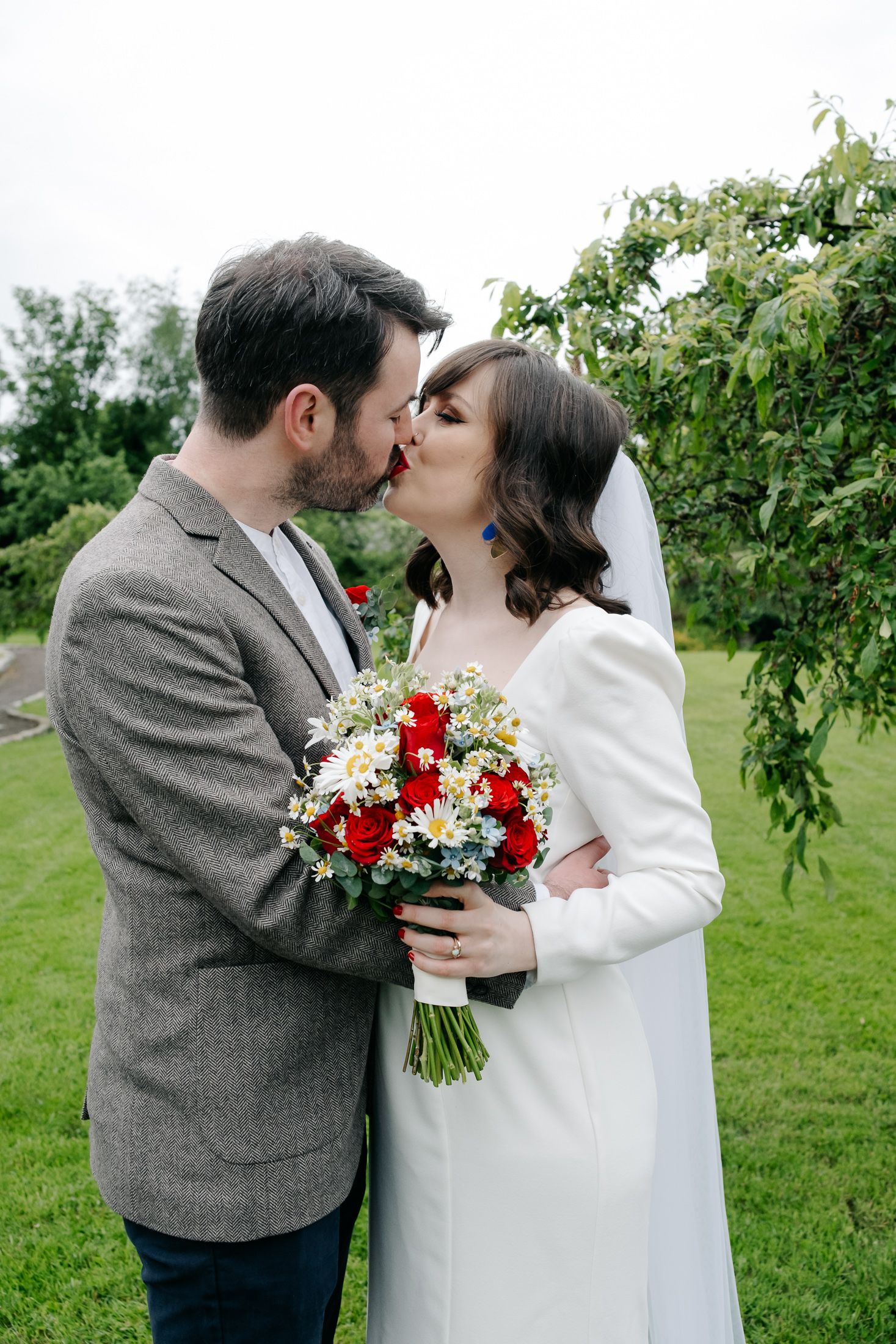 Bride and groom share a kiss outdoors, bride holding red roses and daisy bouquet, groom in gray tweed jacket.