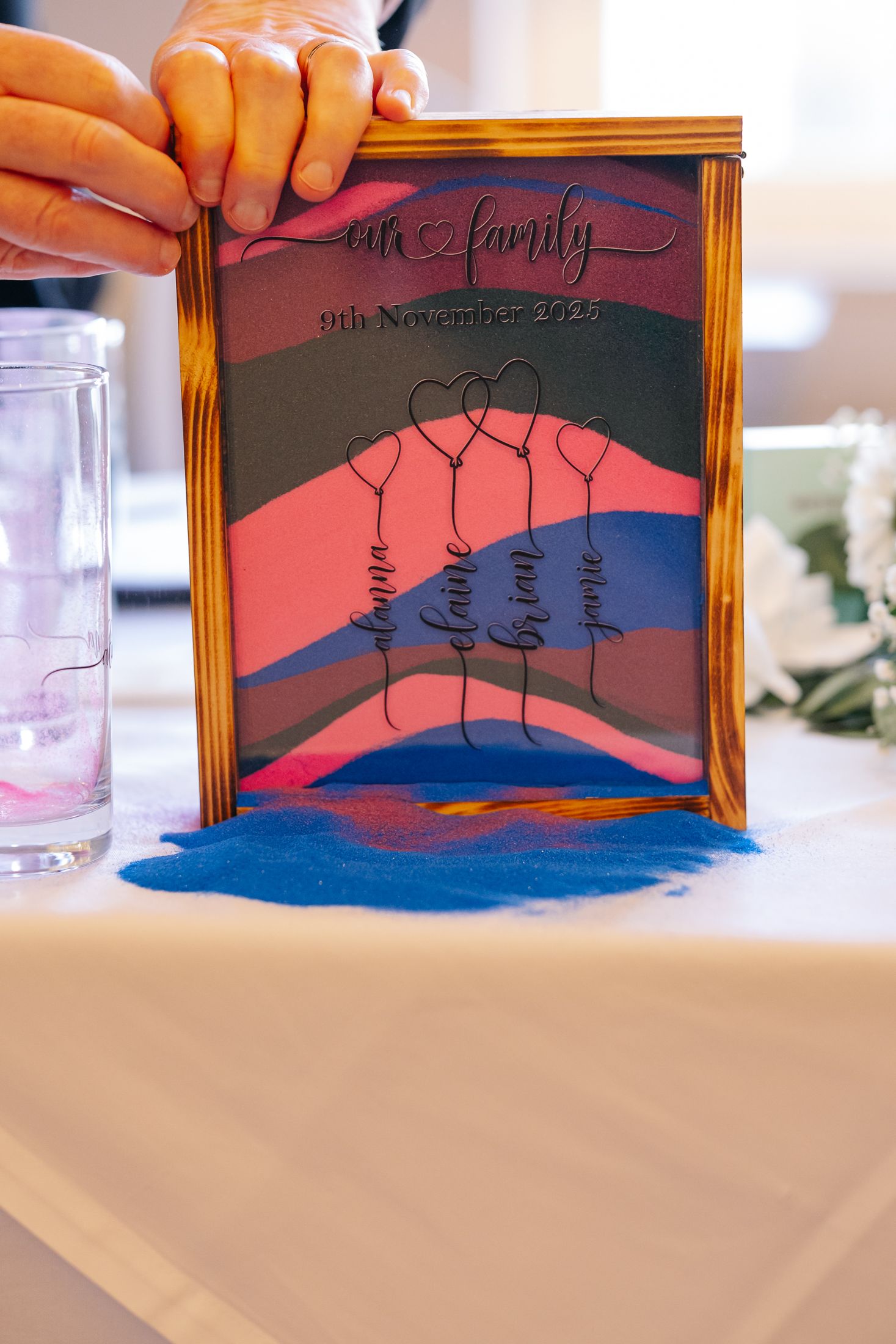 Family sand ceremony frame dated 9th November 2023 with colorful layered sand and wire family member names on display.