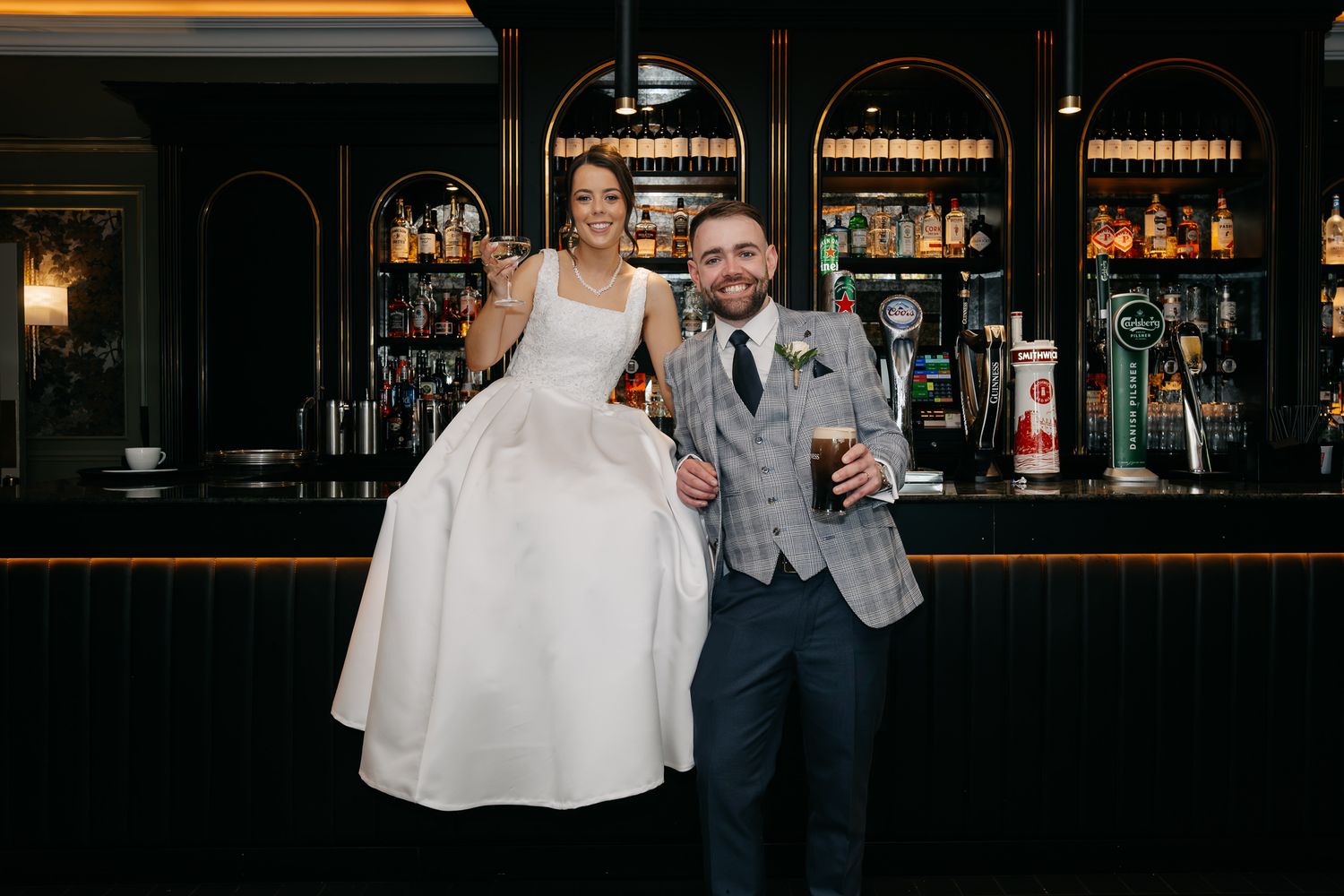 Bride and groom posing happily at a stylish bar, bride seated on counter in white gown, groom holding a drink.