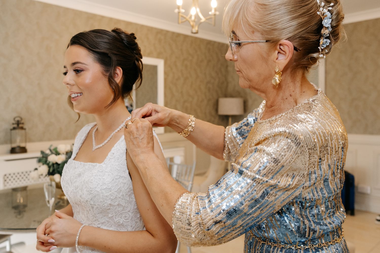 Mother helping bride with pearl necklace in elegant dressing room before wedding ceremony.