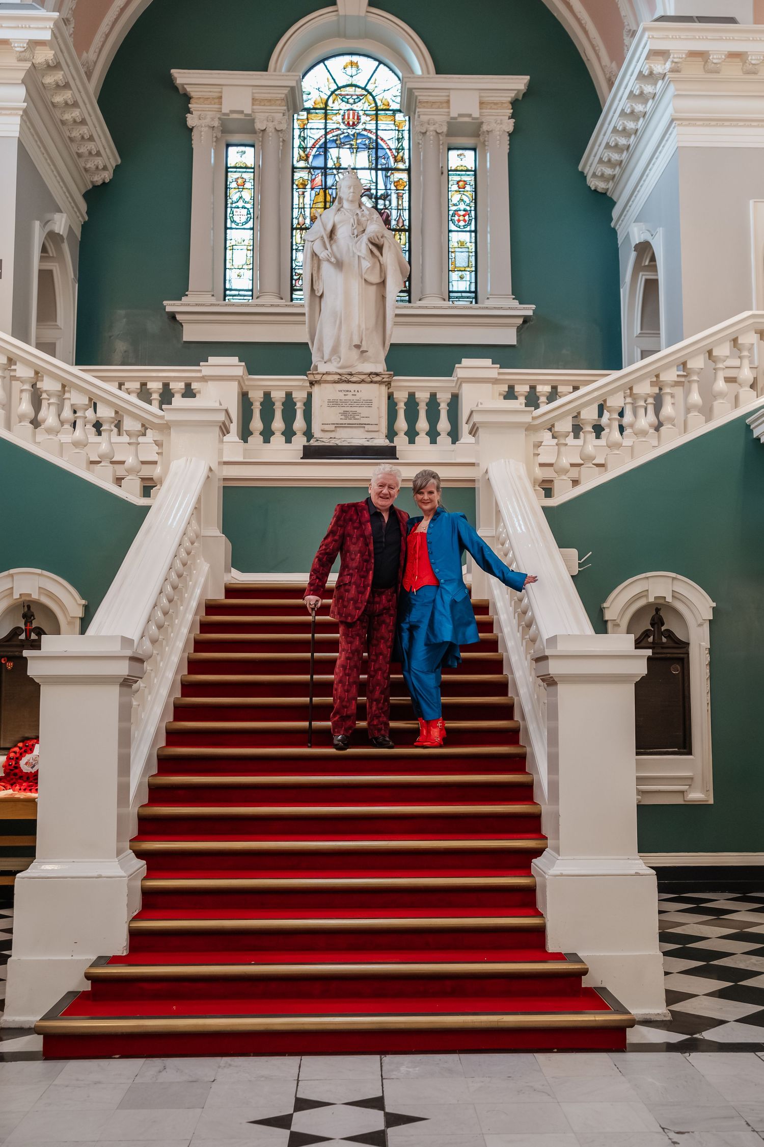 Two people in bright formal attire descending grand staircase with stained glass window.