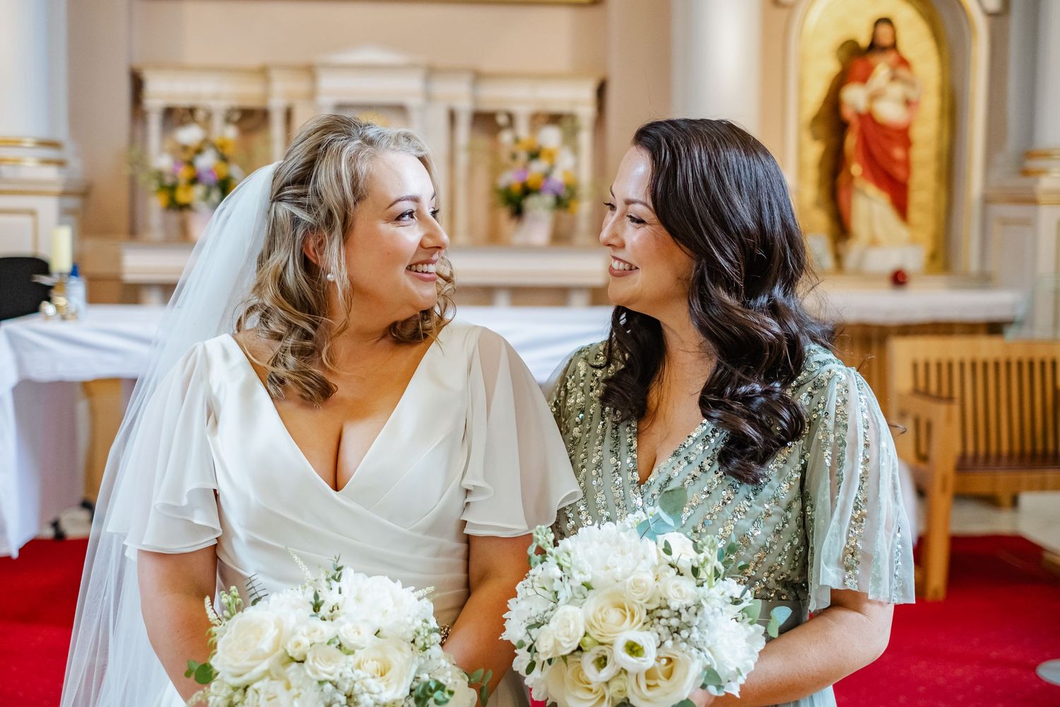 Two women in wedding attire share joyful moment while holding white bouquets in church.