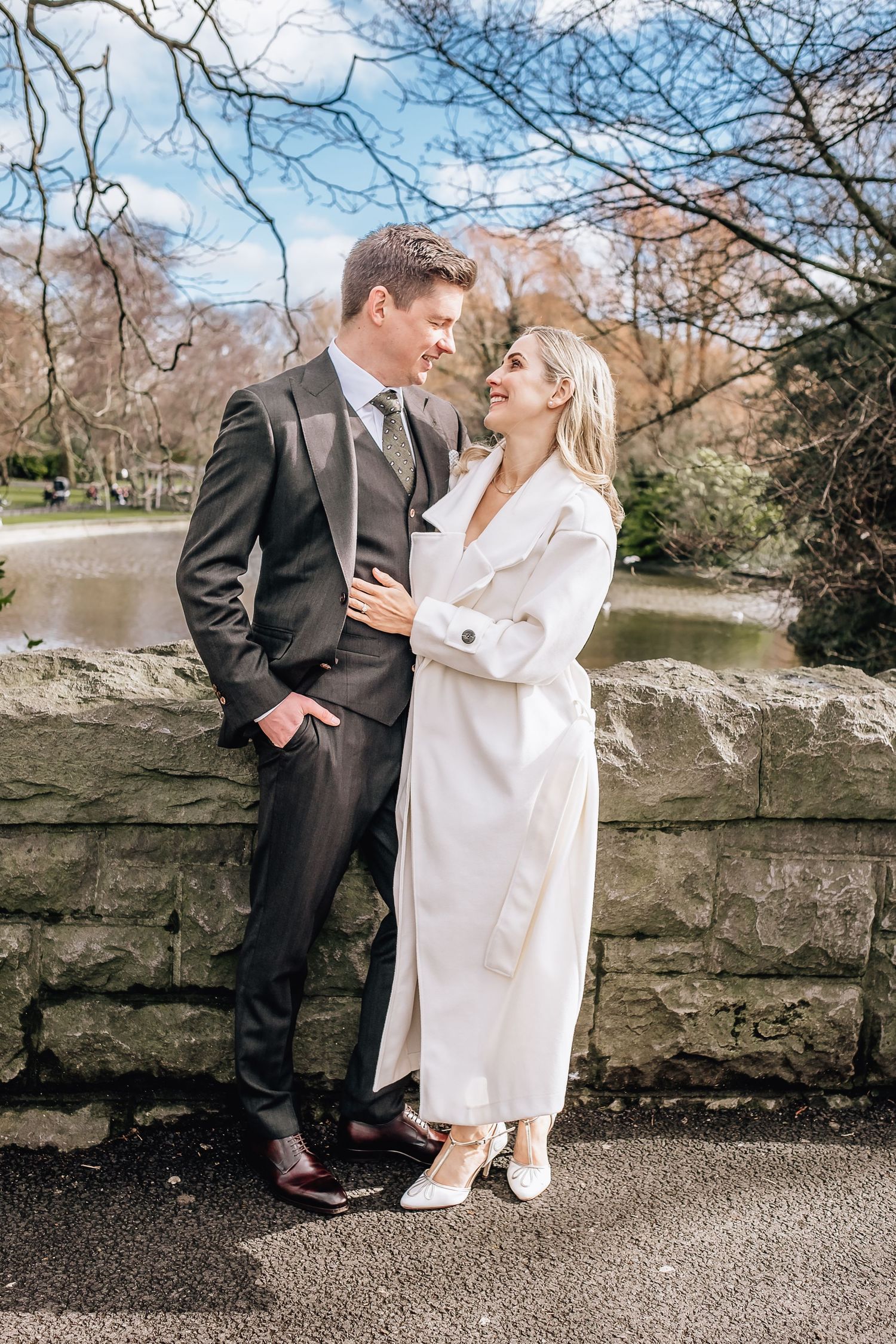 Couple in formal attire share romantic moment by stone wall overlooking pond.