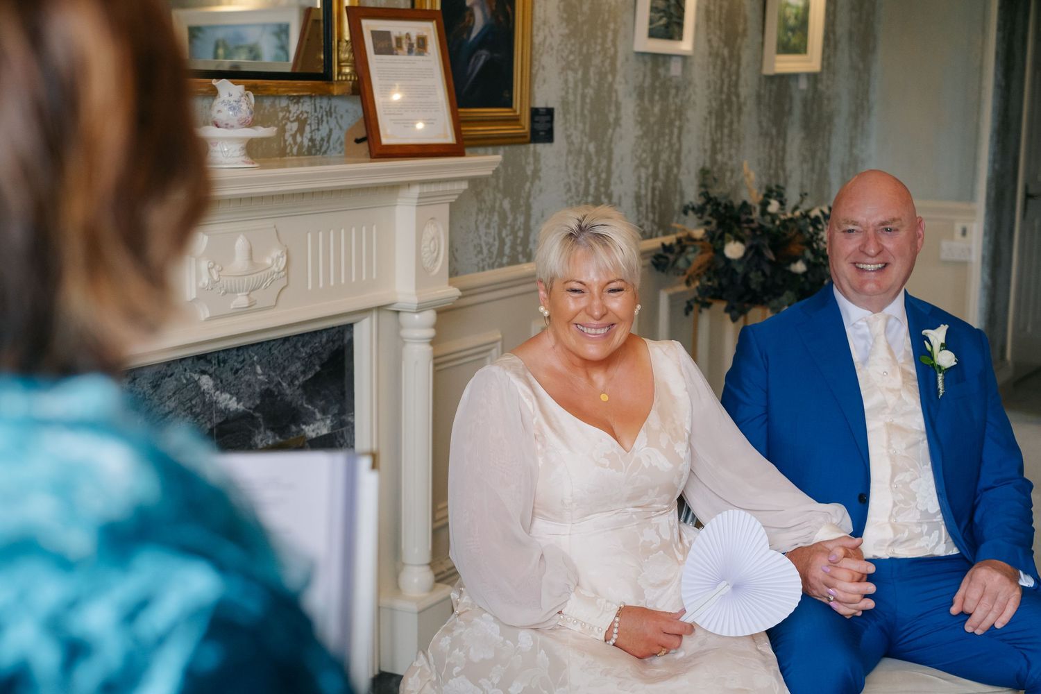 Smiling bride in white gown holding fan seated beside bald groom in blue suit at wedding.