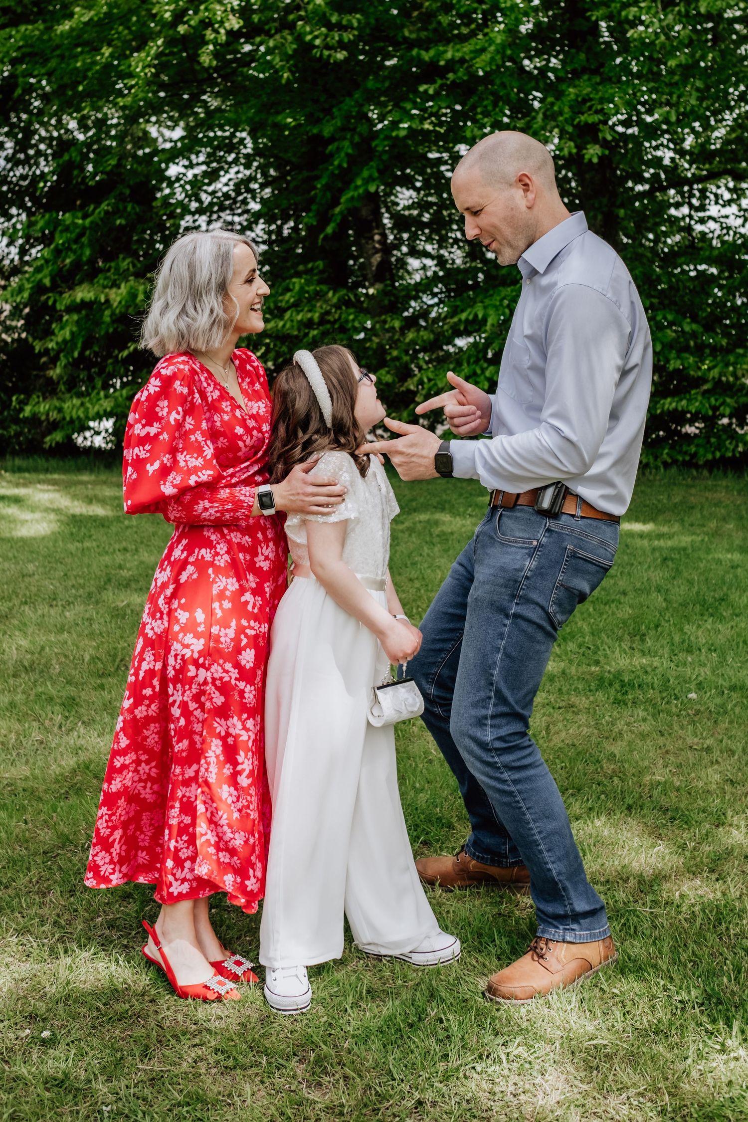 A family of three smiling outdoors, with a woman in red floral dress, girl in white, and man in blue jeans.