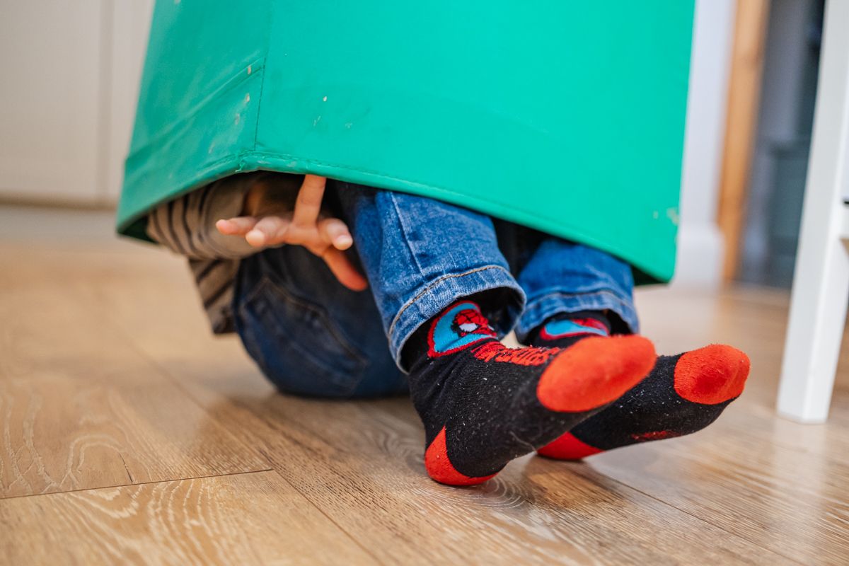Child hiding under a green bucket on hardwood floor wearing colorful Spiderman socks and blue jeans.
