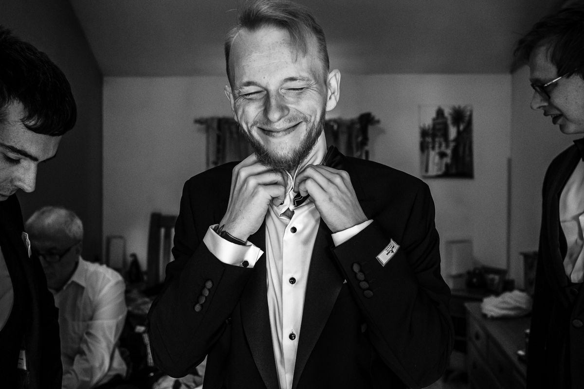 A black and white photo of someone adjusting their bowtie while smiling during formal preparations.