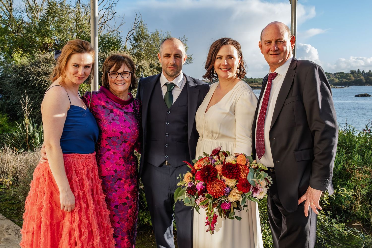 Wedding couple with family posing outdoors near waterfront, bride holding vibrant autumn bouquet.