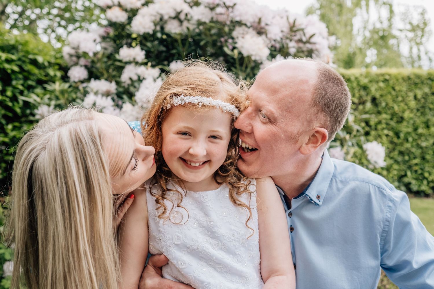 A joyful family portrait with parents kissing their smiling young daughter outdoors near blooming white flowers.