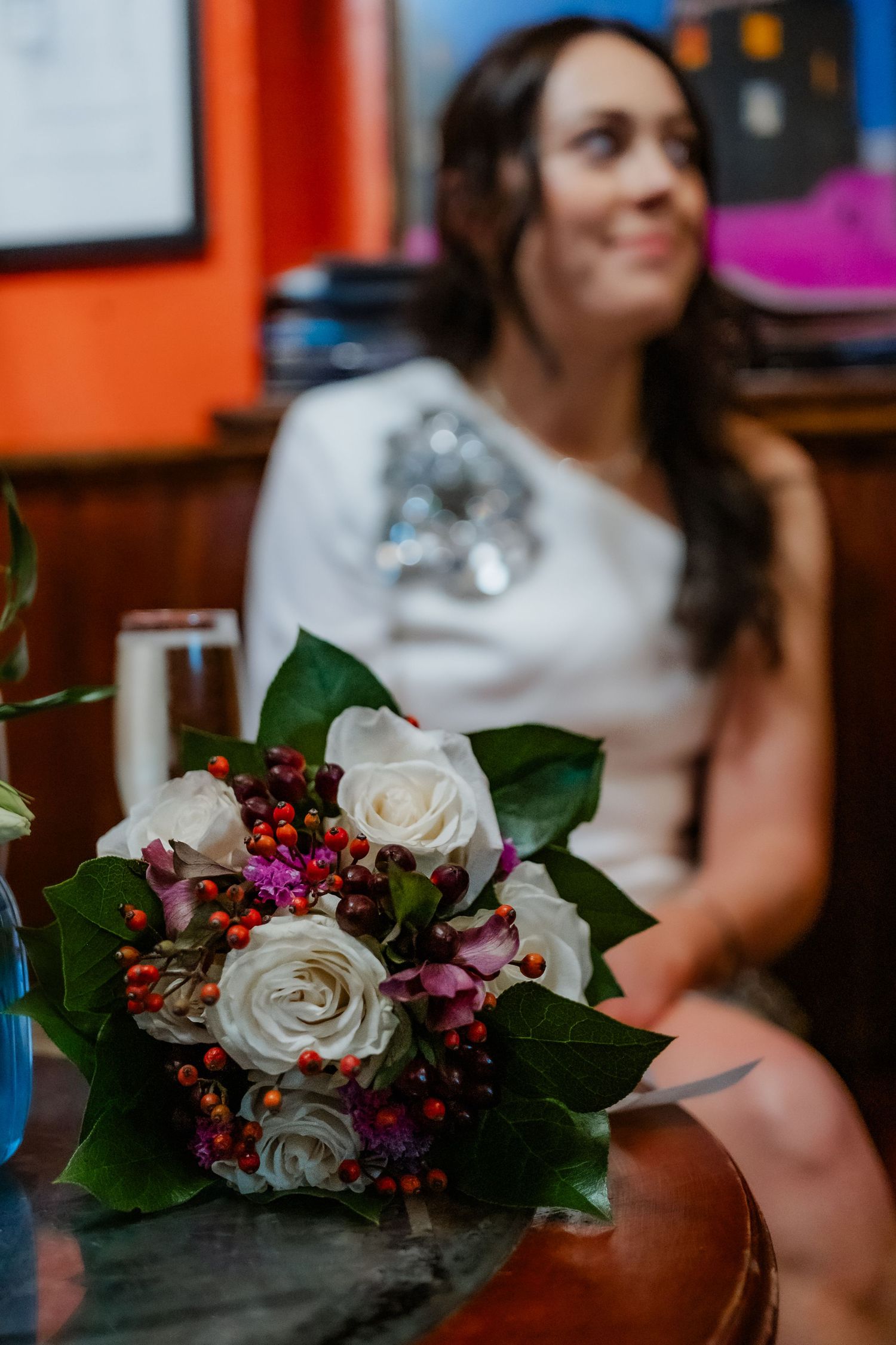 Elegant bridal bouquet of white roses and red berries on a table with smiling bride in white dress in background.