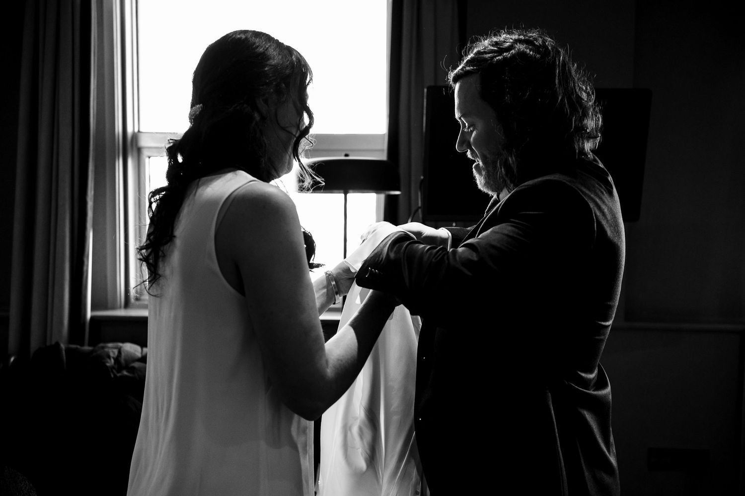 Black and white photo of a bride being helped into her wedding dress by a woman near a bright window.