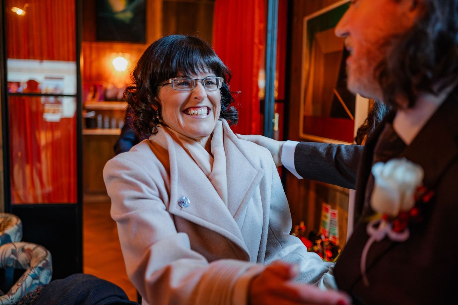 Smiling woman in white coat at wedding event holding hands with man wearing boutonniere in warmly lit venue.