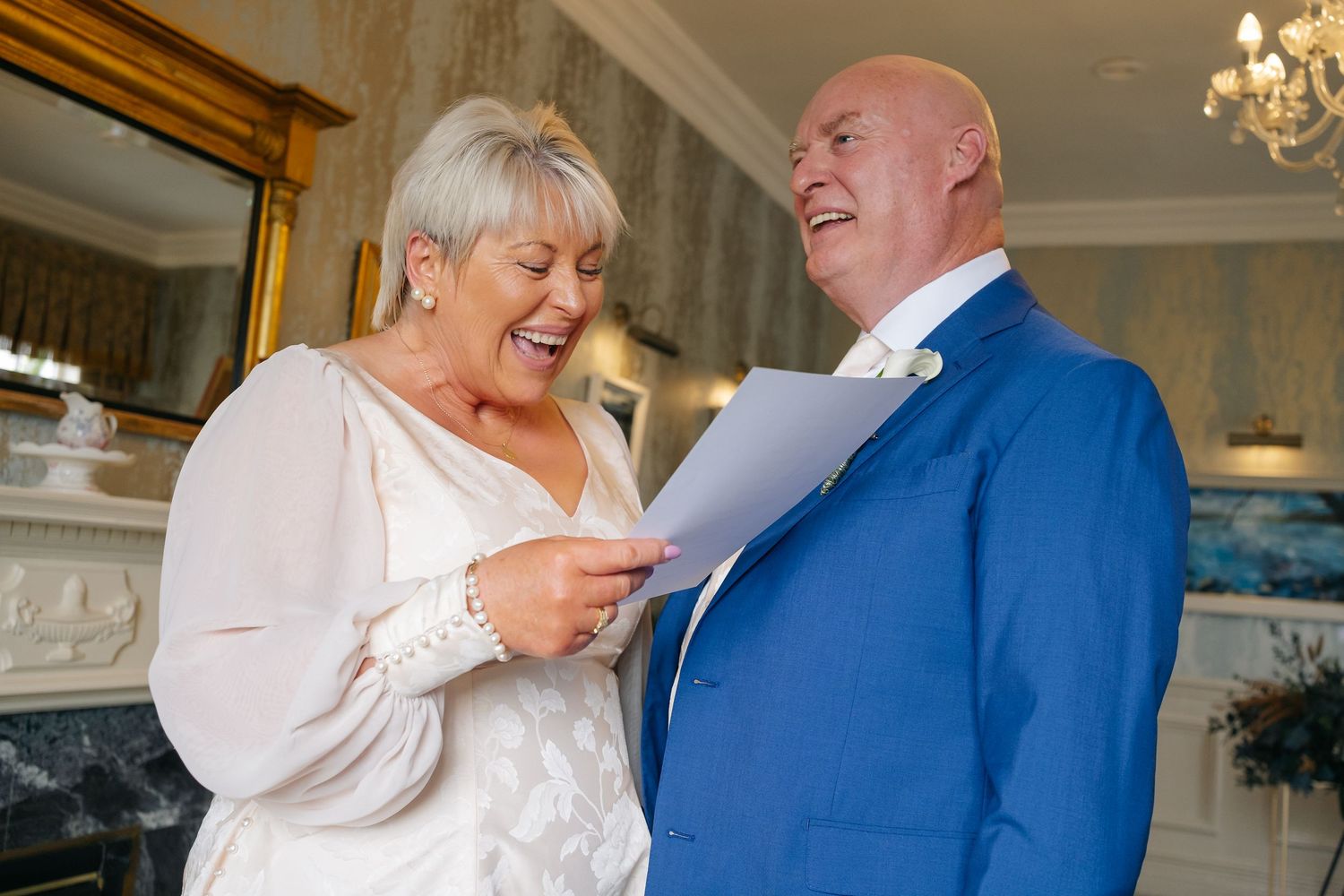 An older bride and groom laugh together while reviewing wedding vows, groom wearing a blue suit.