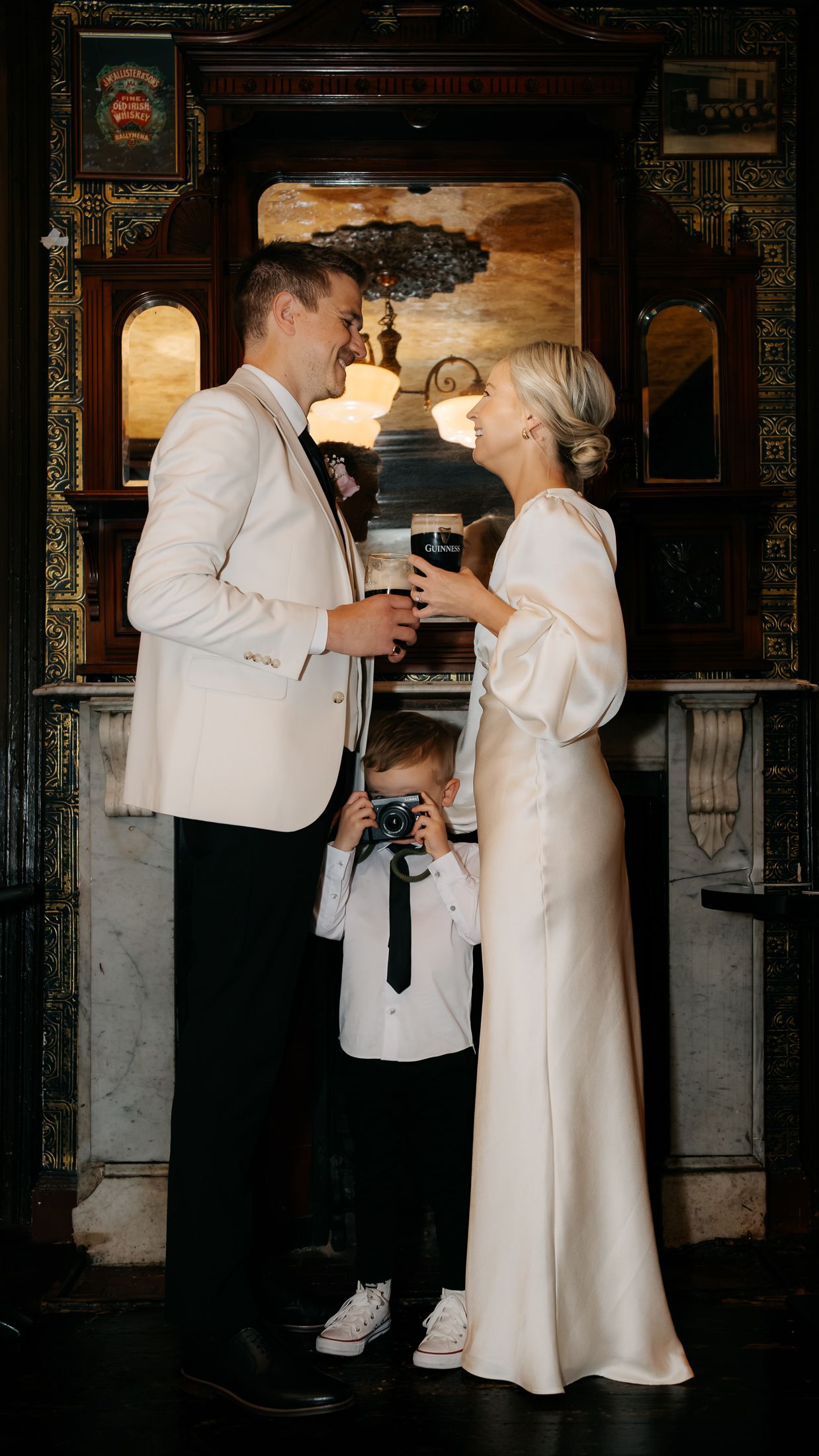 Couple in wedding attire toasting drinks while a young boy photographs them in an elegant venue.