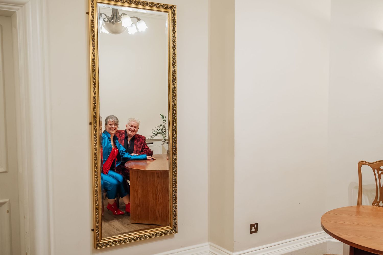 Reflection in gold-framed mirror shows blue and red outfits in interior space.