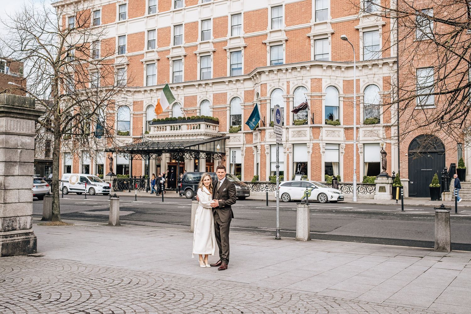 A couple stands together outside the elegant facade of The Shelbourne Hotel in Dublin on a winter day.