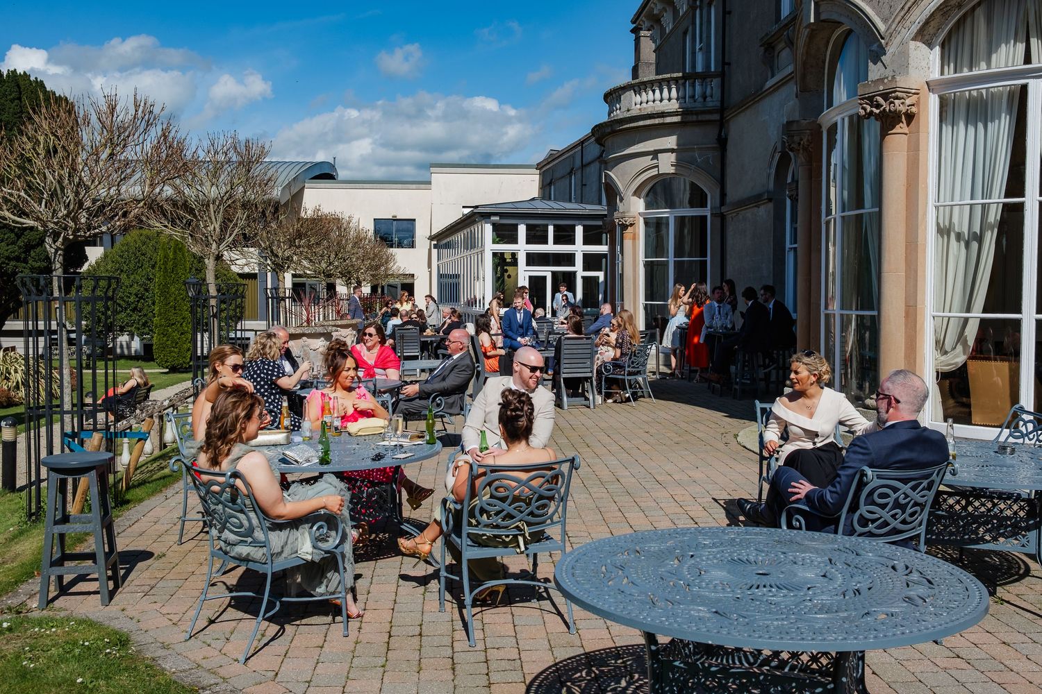 Diners enjoy outdoor seating at restaurant tables beneath blue skies and white clouds on a sunny day.