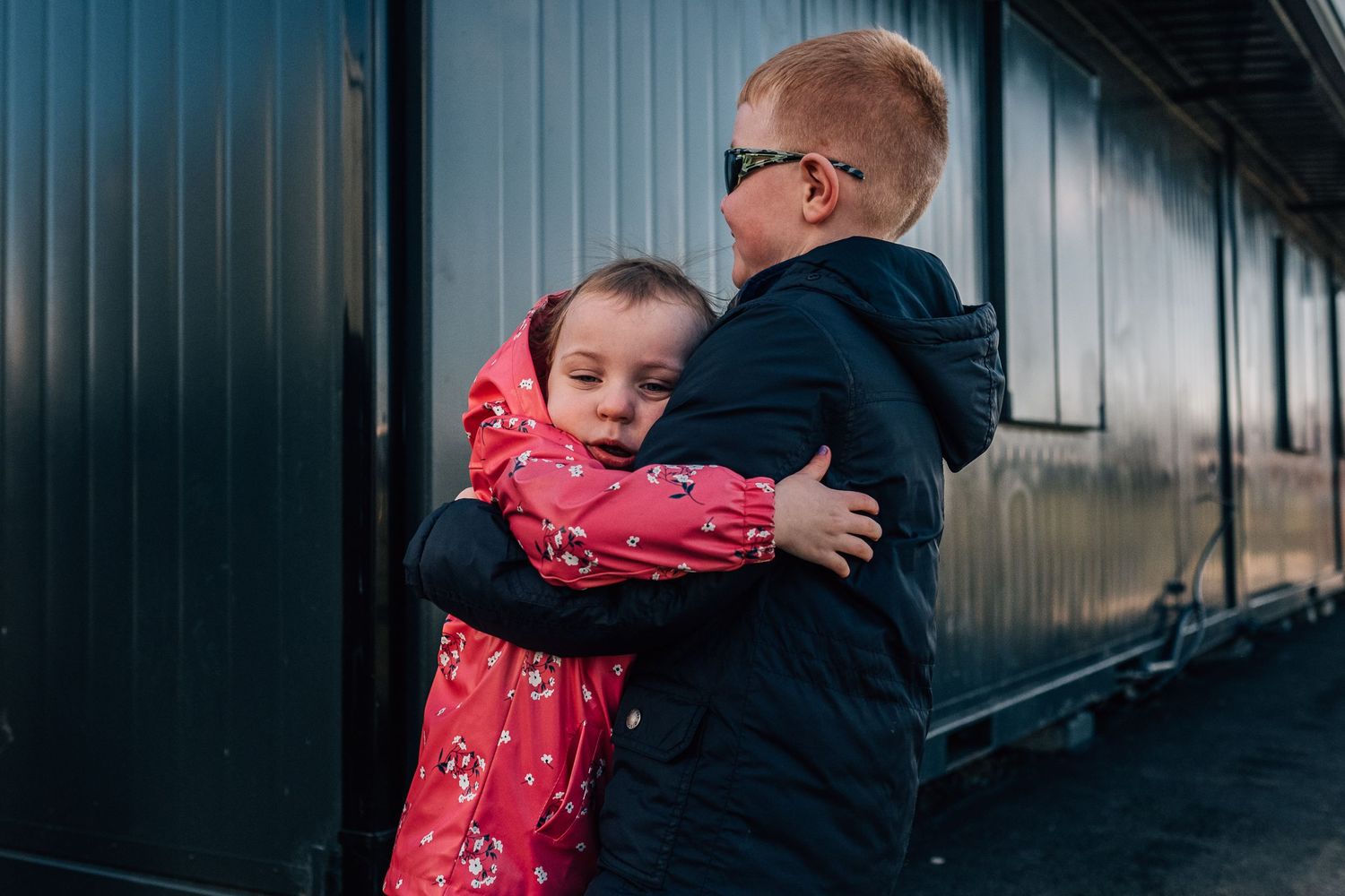 Two children sharing an embrace against a dark metal wall outdoors.