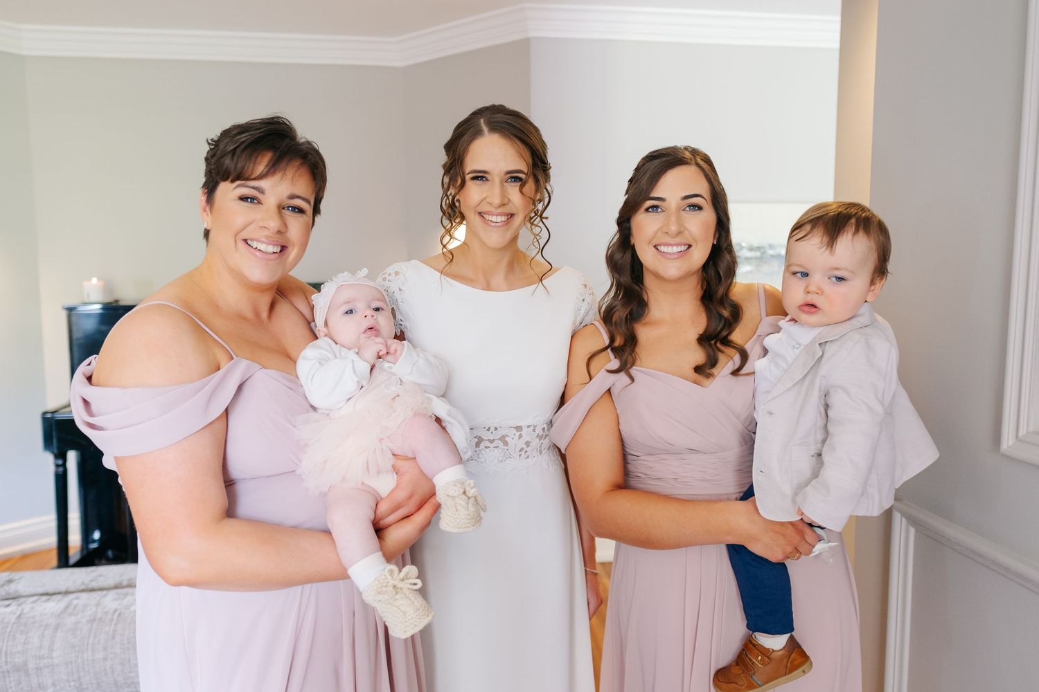 A group wearing pale pink dresses and holding infants stands together in a light-colored interior space.