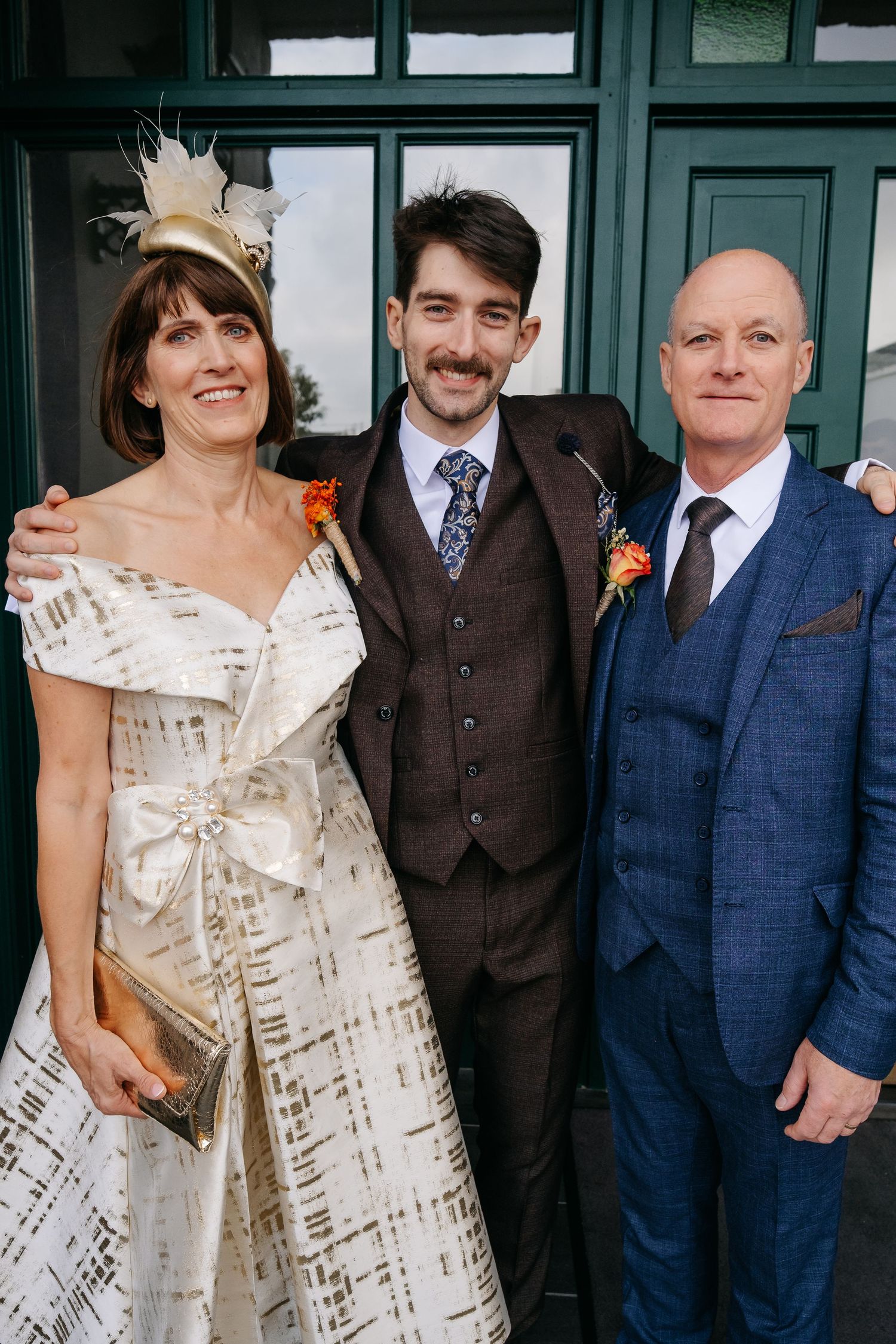 Wedding group stands together wearing navy suits and cream-colored dresses against green doors.