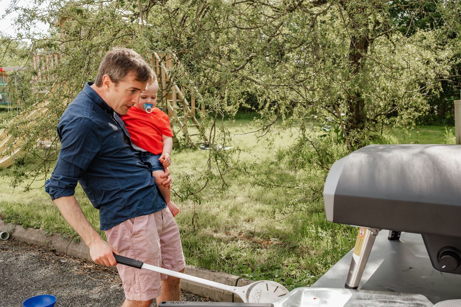 Person grilling outdoors while holding small child in red clothing.